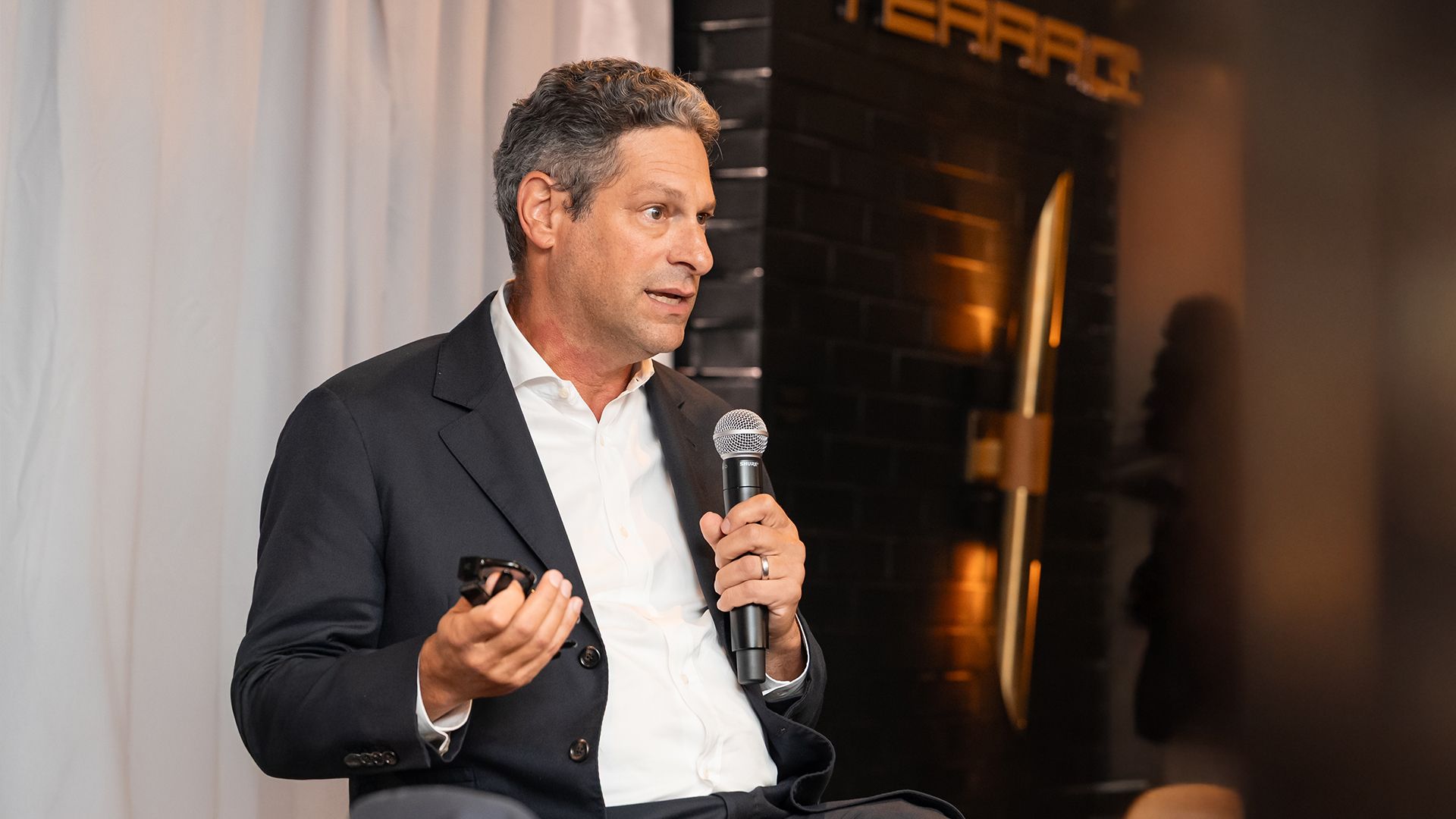 Man in a dark suit and white shirt speaking into a microphone, gesturing with his other hand, sitting against a backdrop with white curtains and dim lighting.