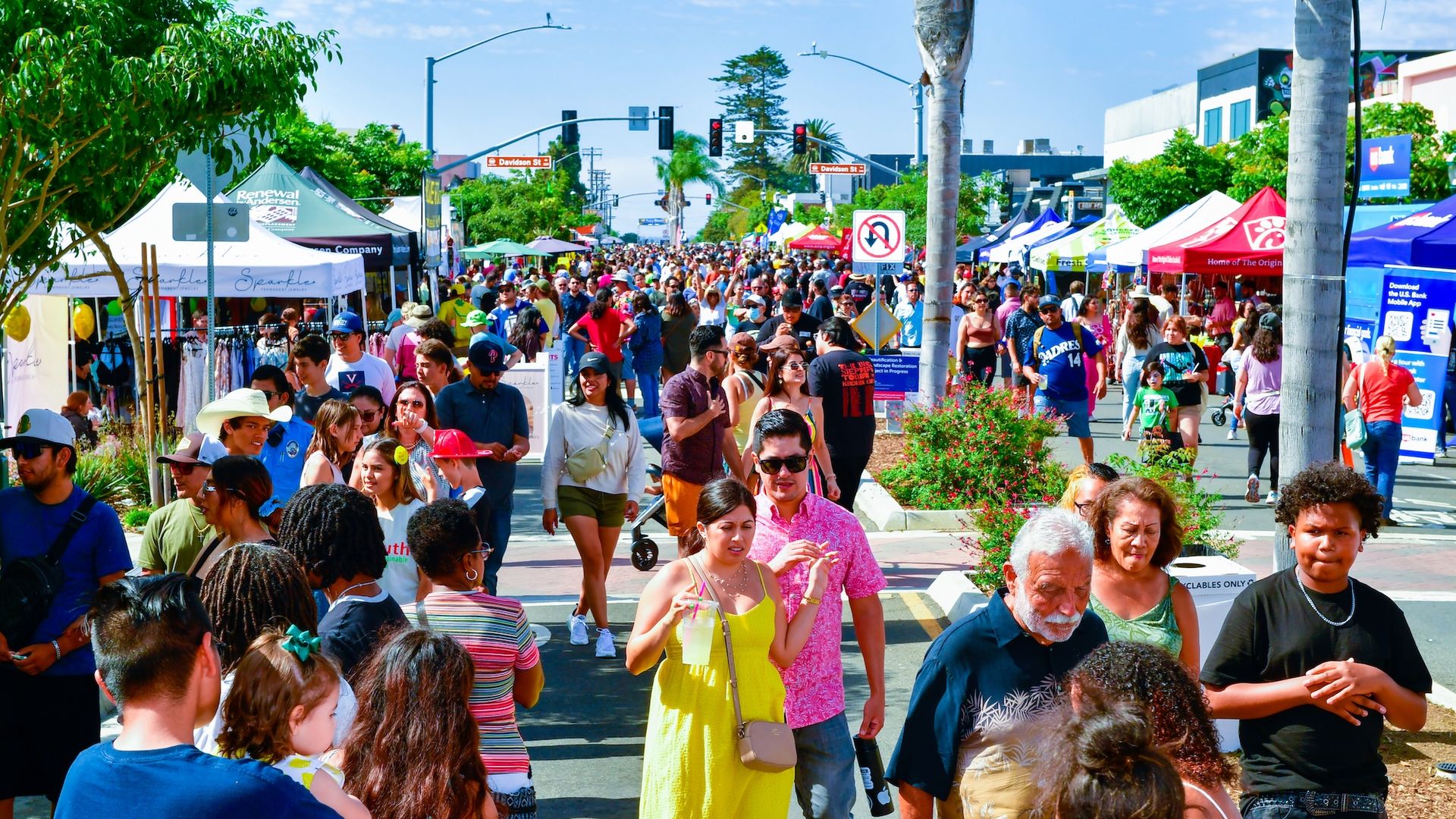 Crowded outdoor festival with many people walking among colorful vendor tents under bright sunny sky, trees and street signs visible in background.