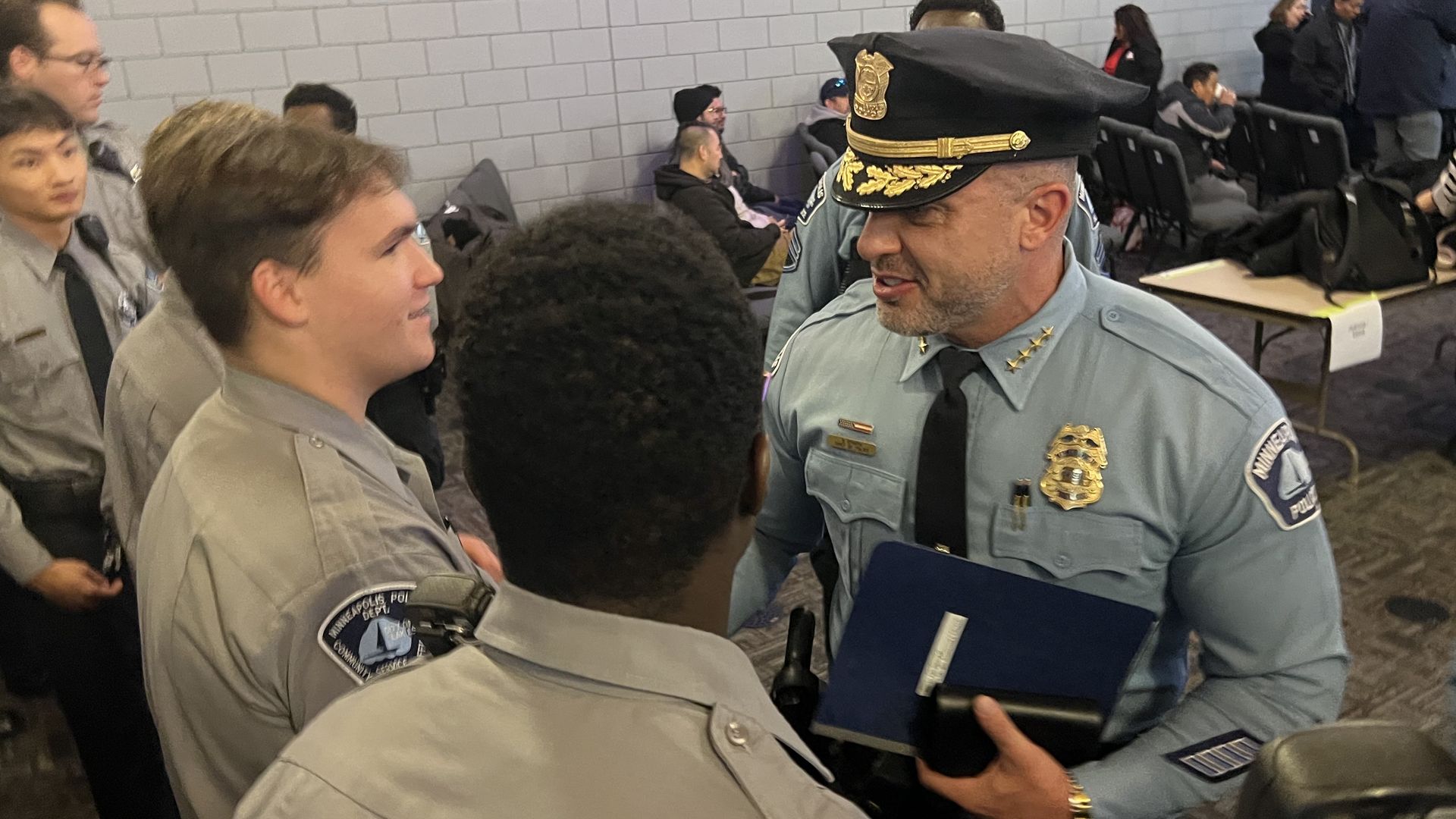 A police captain in a light-blue uniform and peaked cap talks with young cadets in gray shirts; he holds a blue binder as others stand and sit in a conference room.