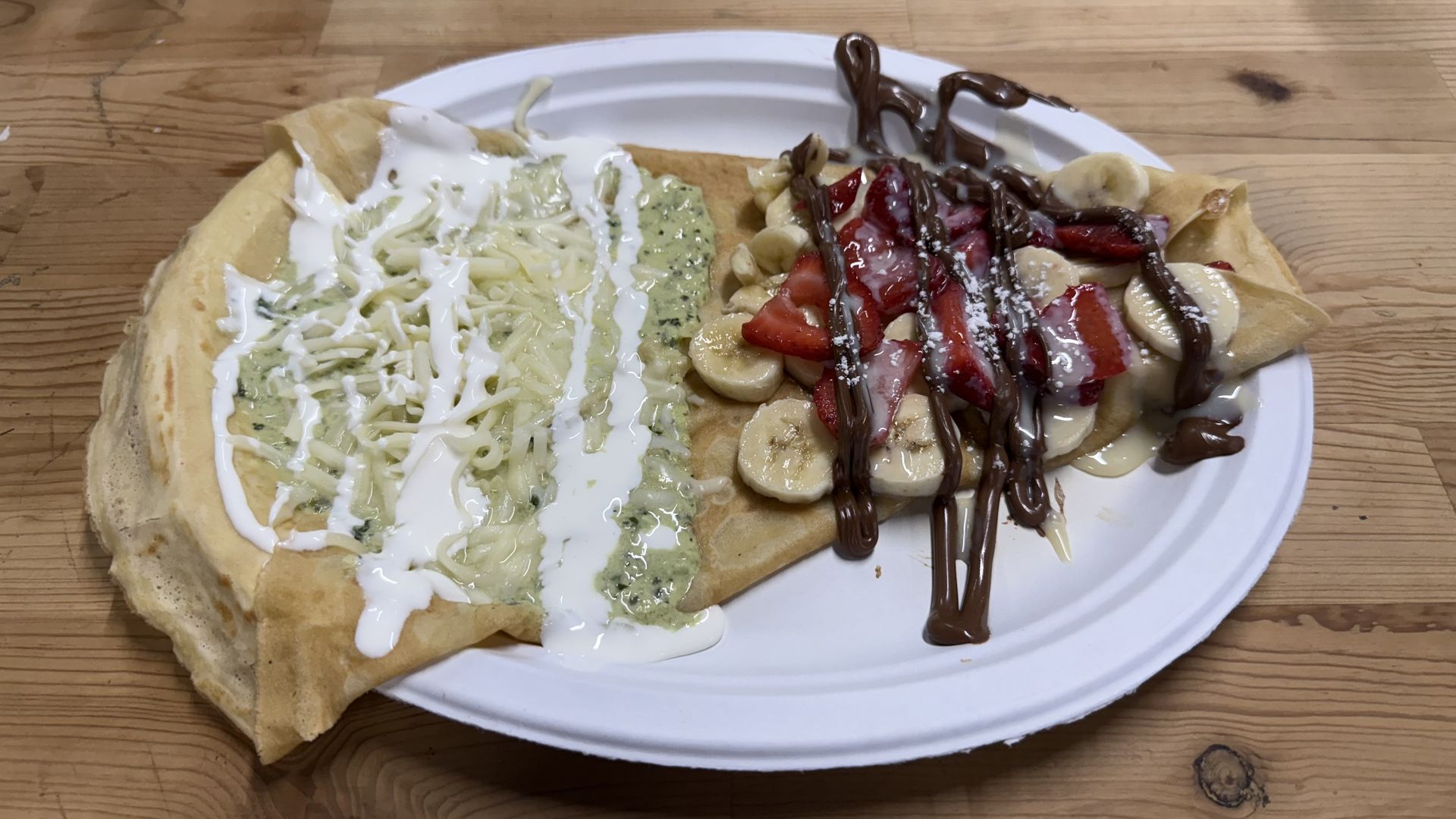 An open-faced, wedge-shaped crepe with green sauce and cheese with a white sauce drizzled on one end, and strawberries and banana slices drizzled with nutella on the other end.