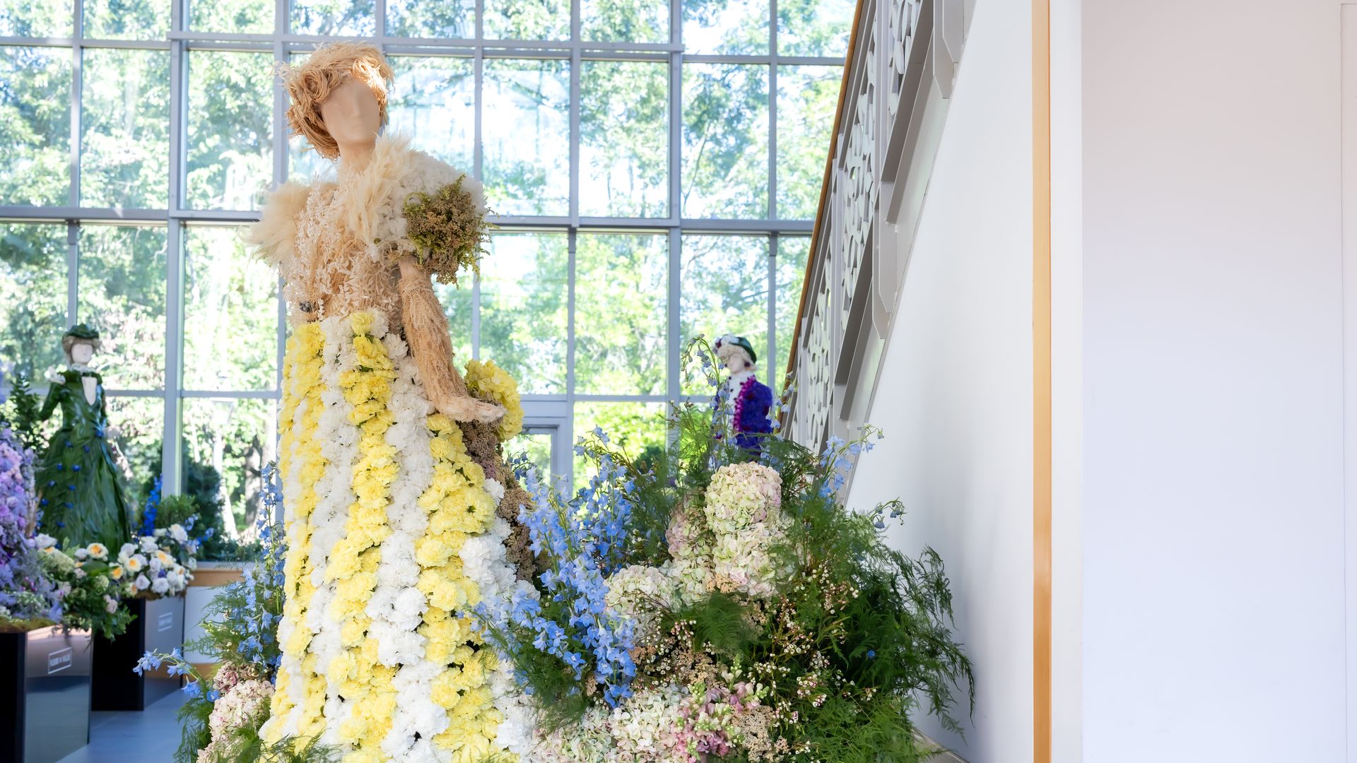 In a sunlit atrium, a tall flower-covered sculpture resembling a dress stands center, layered in yellow and white blooms with greenery, near a modern lattice staircase.
