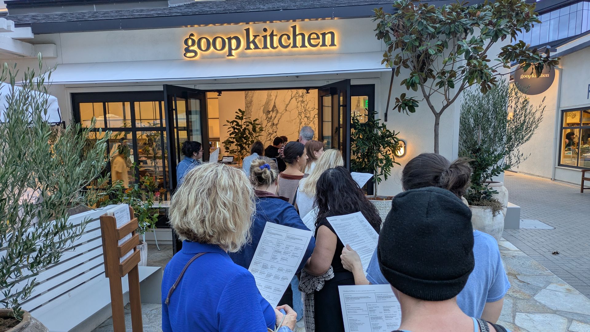 Customers queue outside Goop Kitchen, a modern restaurant with a glowing sign. Open glass doors reveal a marble-walled interior, surrounded by potted trees and greenery.
