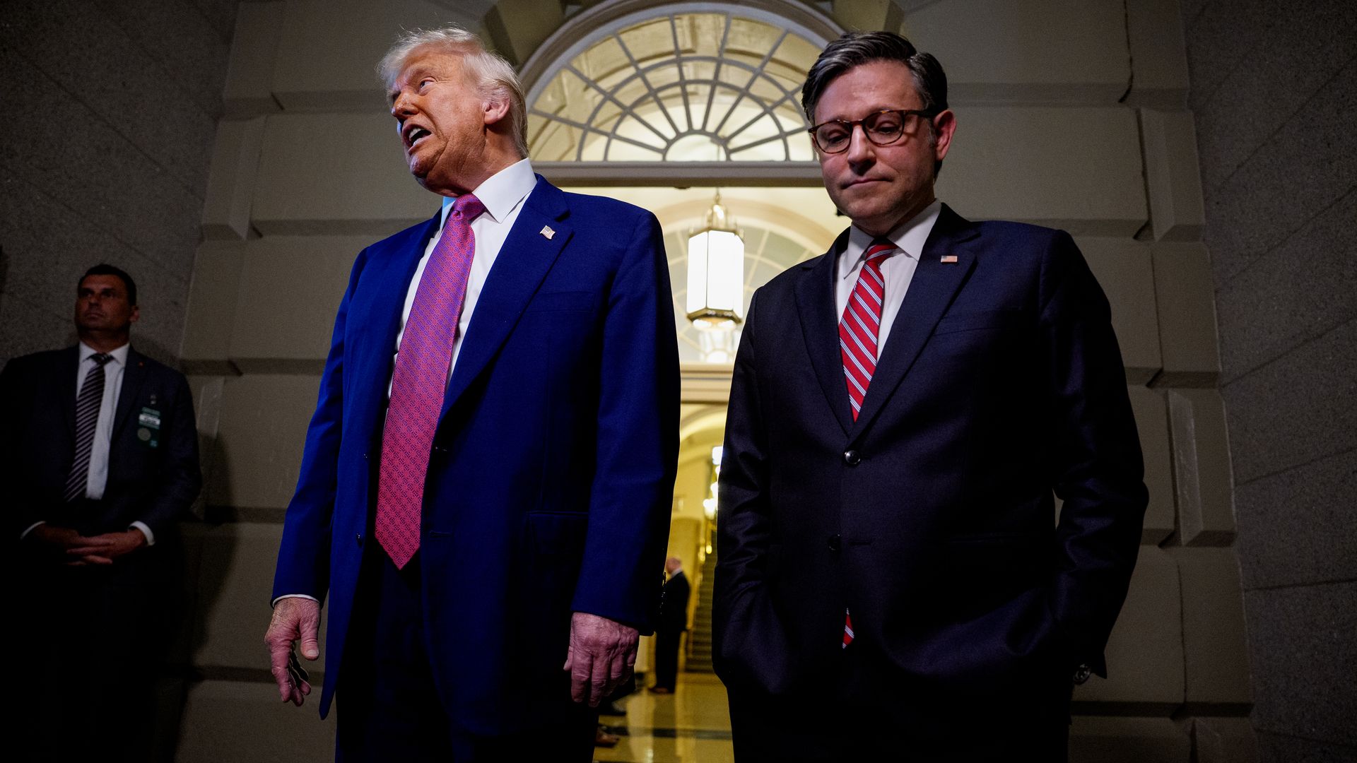 Donald Trump and Mike Johnson, both wearing blue suits, stand in a sandstone hallway while surrounded by Secret Service agents.