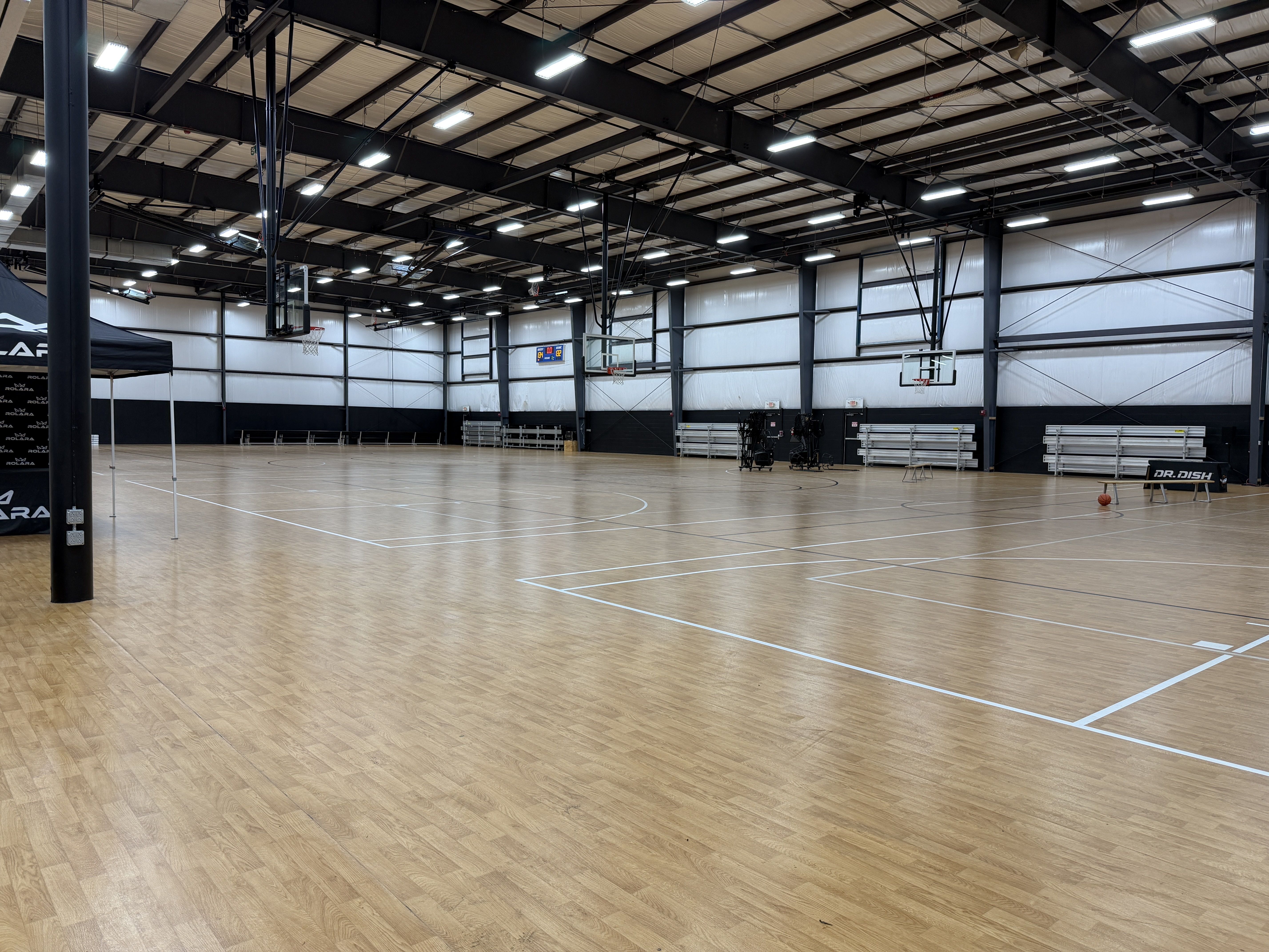 An empty indoor basketball gym with a polished wooden floor, multiple hoops and backboards, high black steel ceiling trusses, bright overhead lights, and folding bleachers along the grey walls.
