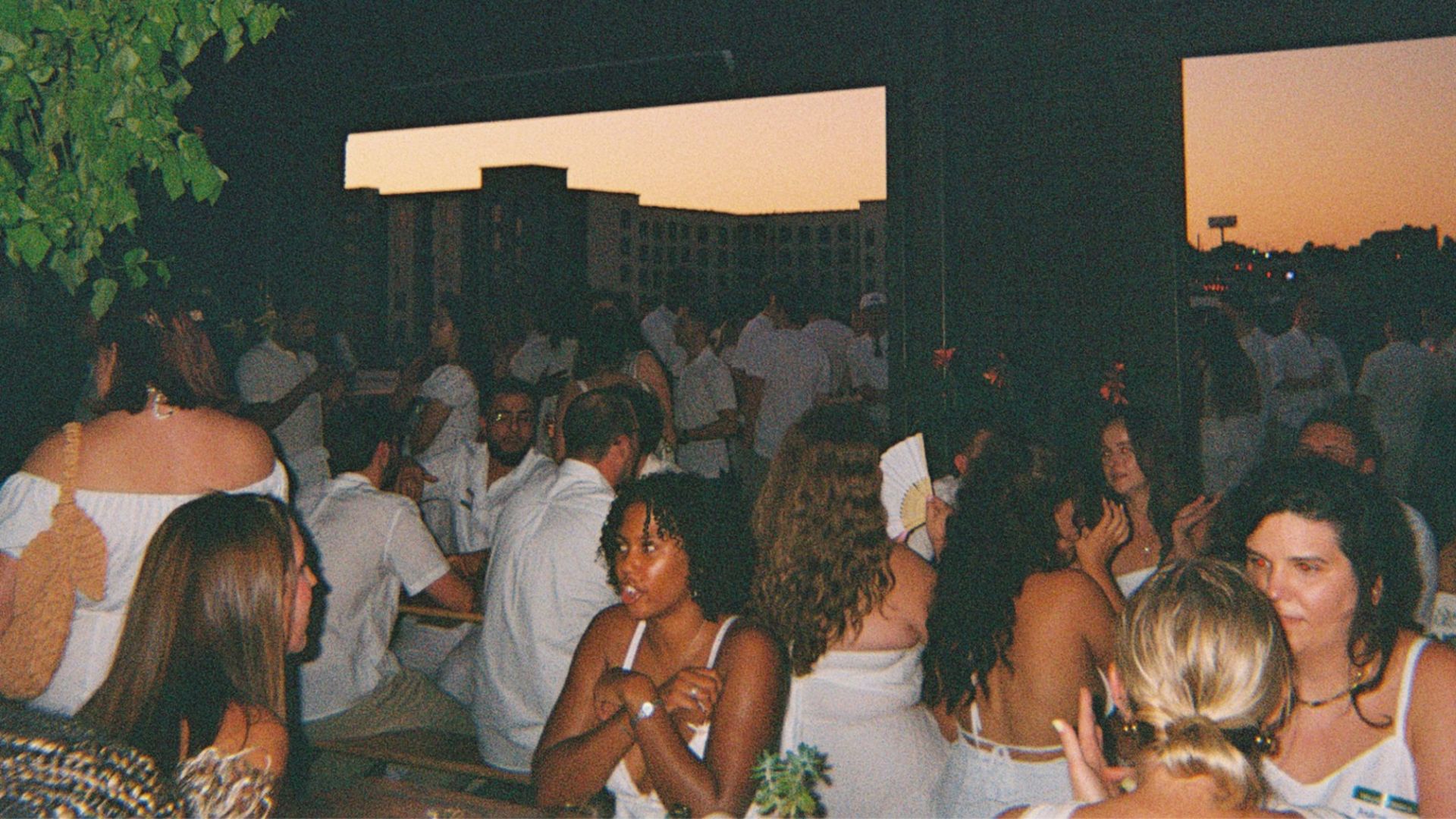 Outdoor party at dusk with many people dressed in white, mingling under a dark overhang. An orange sunset glows through large openings, and a leafy plant hangs to the left.