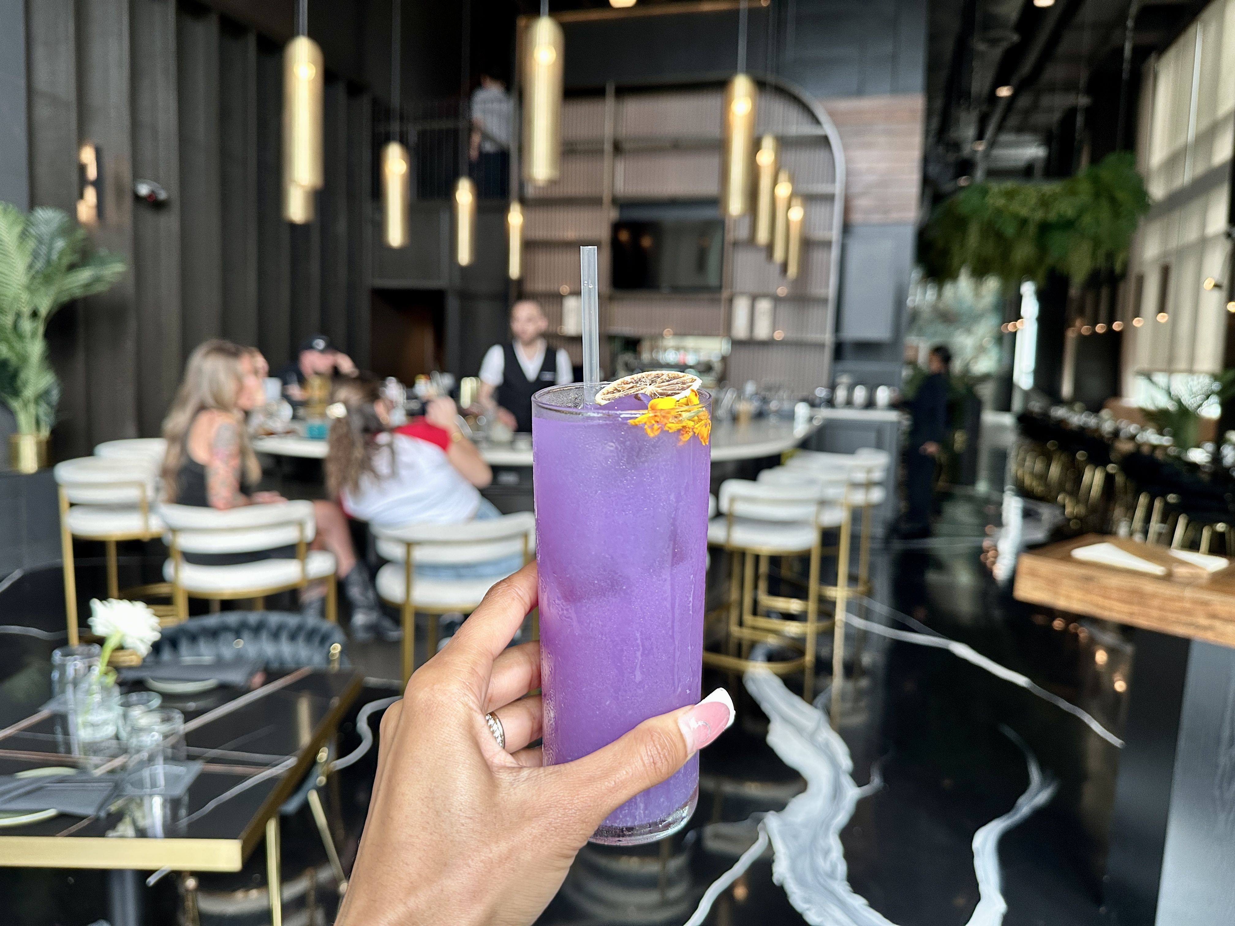 A purple cocktail with a dried lemon garnish is held by a hand with pink nails and a ring; a modern restaurant with gold pendant lights and blurred diners in the background.