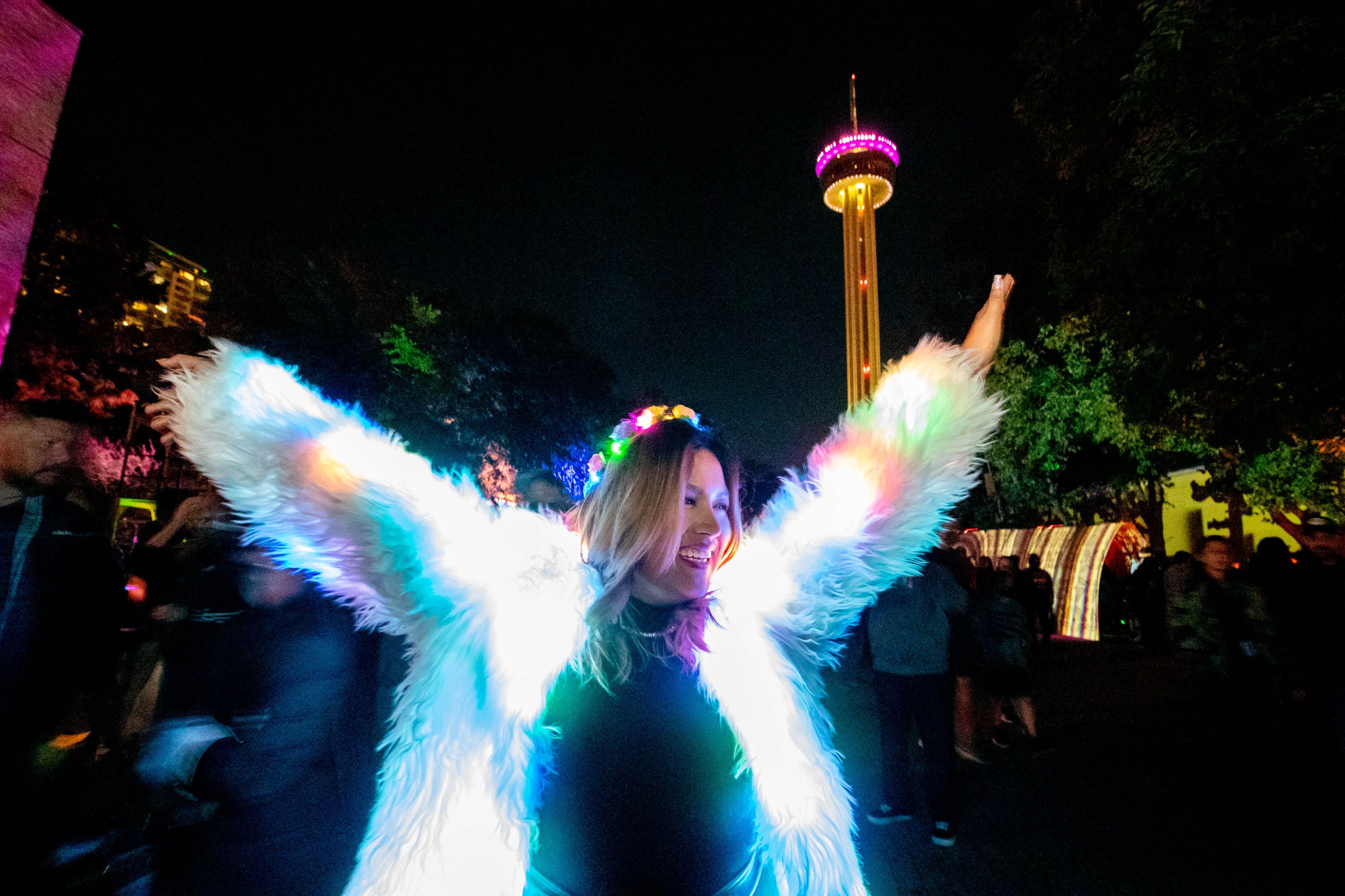 A woman wearing an illuminated jacket and tiara extends her arms across a night sky with the Tower of Americas in the background. 