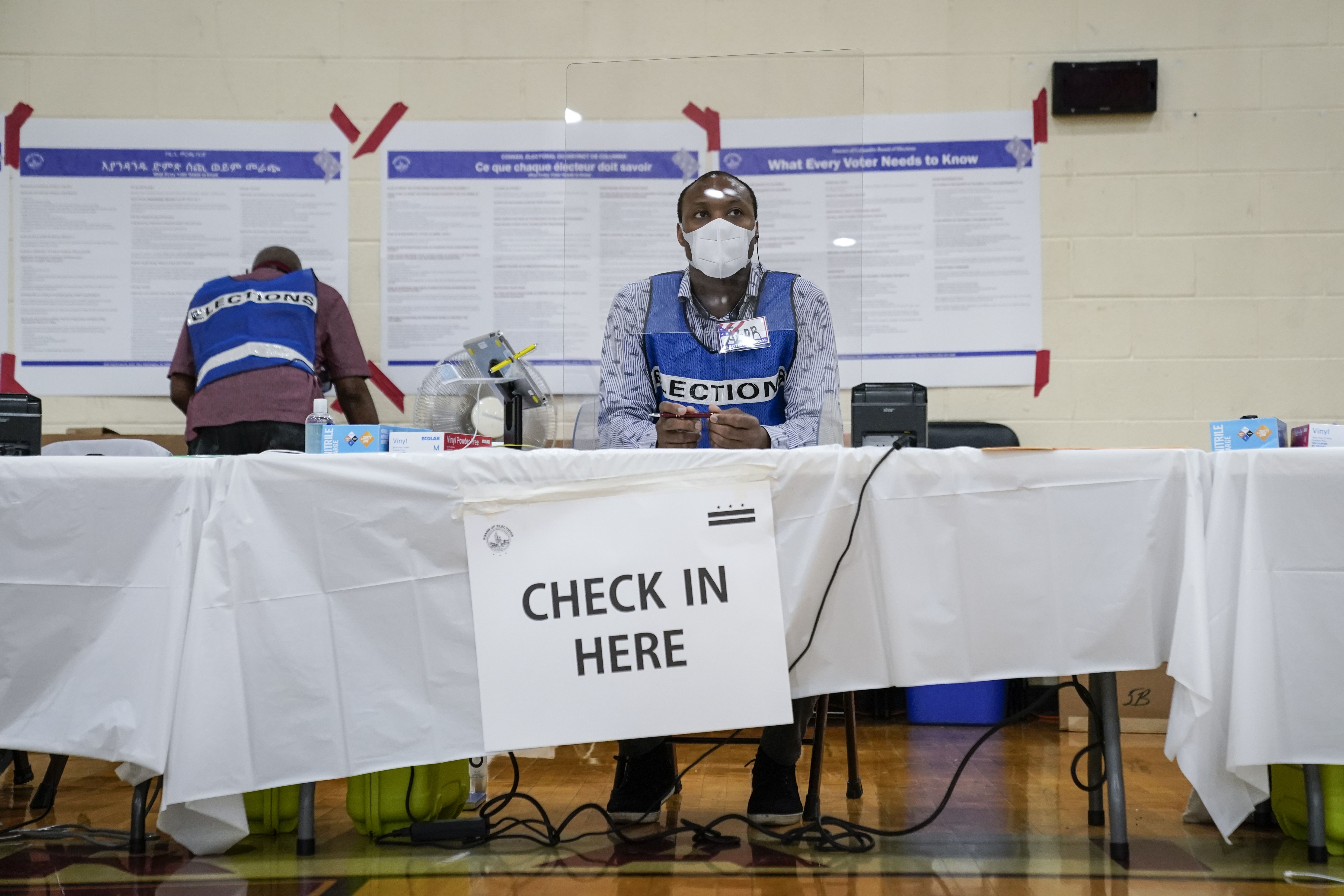A woman sits at a table with a banquet sheet over it while wearing a face mask. A sign on the table reads "check in here"