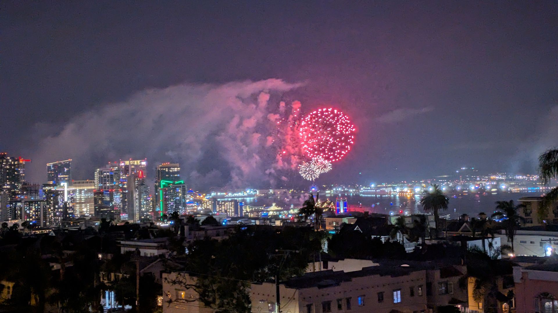 Night-time city skyline with illuminated buildings; pink-red fireworks burst above clouds in a purple-hued sky, lighting the urban landscape with colorful bursts.