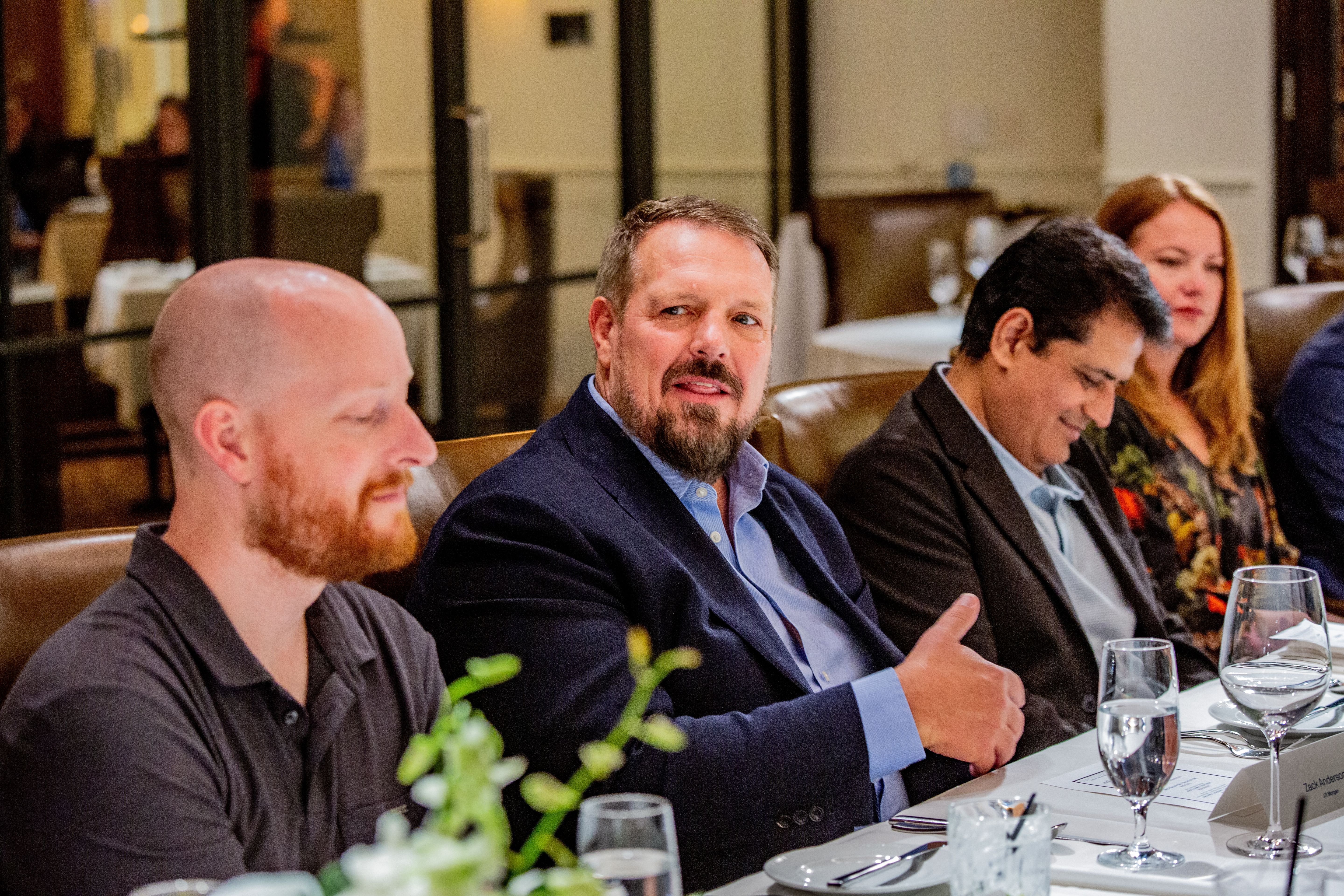 Group of professionals at a formal dinner in a restaurant. Centered man with a beard in a navy blazer speaks; others sit nearby, some looking down. Table set with plates, glasses, and menus.