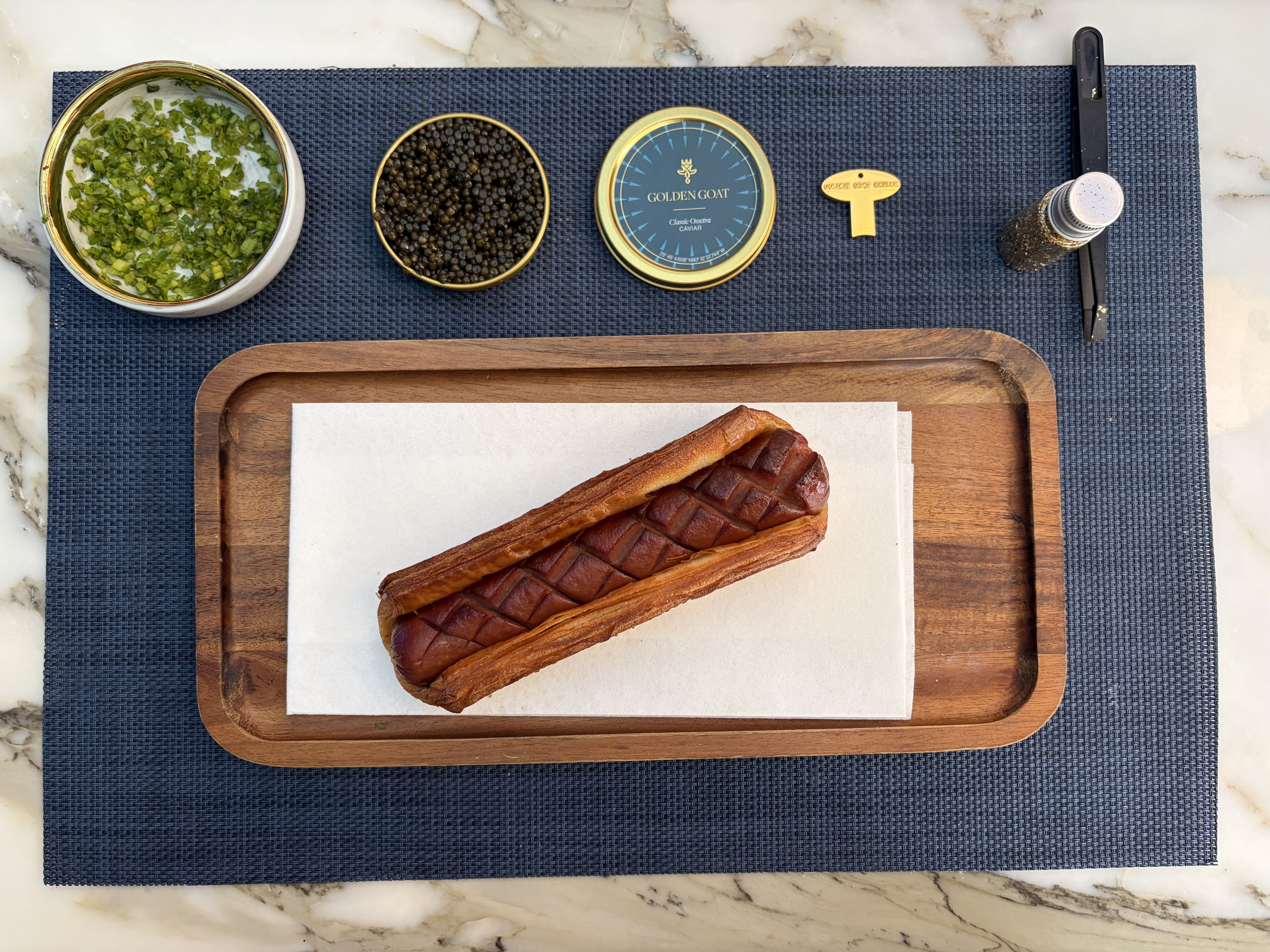 Top view of a hot dog with cross-hatched sausage in a toasted bun on wooden tray with caviar tin, chopped chives, caviar spoon, black pepper grinder, and tweezers on blue mat.