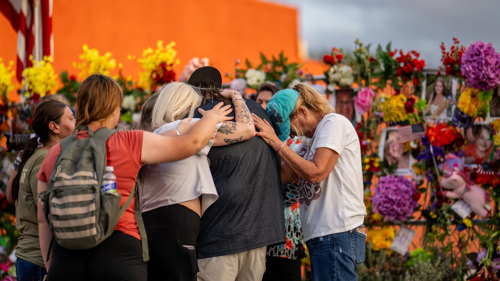Community members gather during a candlelight vigil to honor the lives lost in the July Fourth flooding last year. About five people are seen hugging each other in a group huddle, with a bright orange wall and a memorial with colorful flowers behind them in the background.