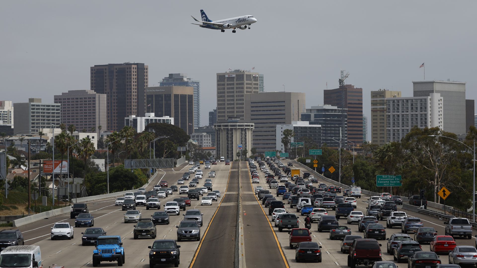 A plane flies over a busy interstate with the San Diego skyline in the background.