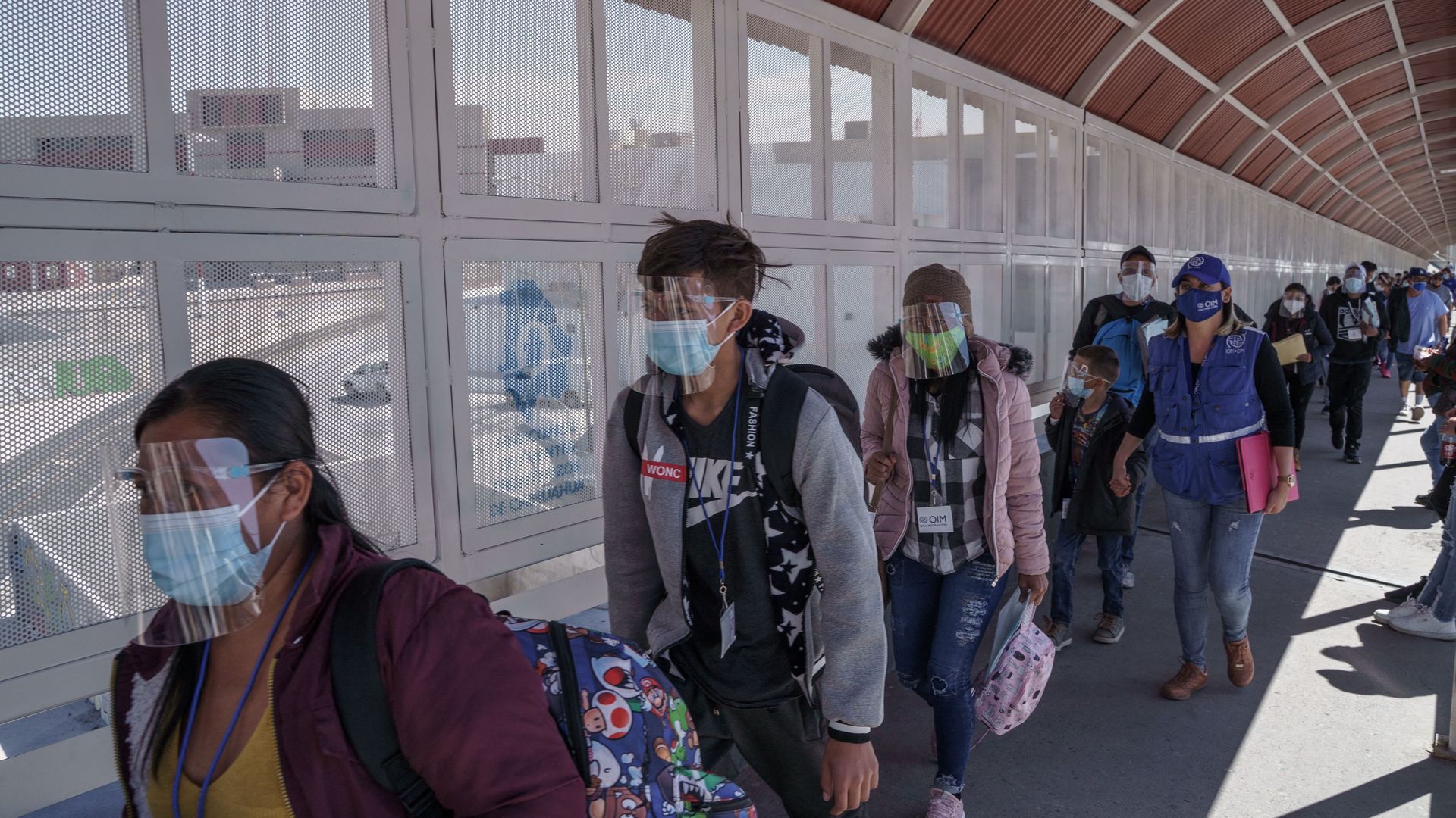 Photo of a group of people wearing masks and carrying luggage in a line waiting on a bridge