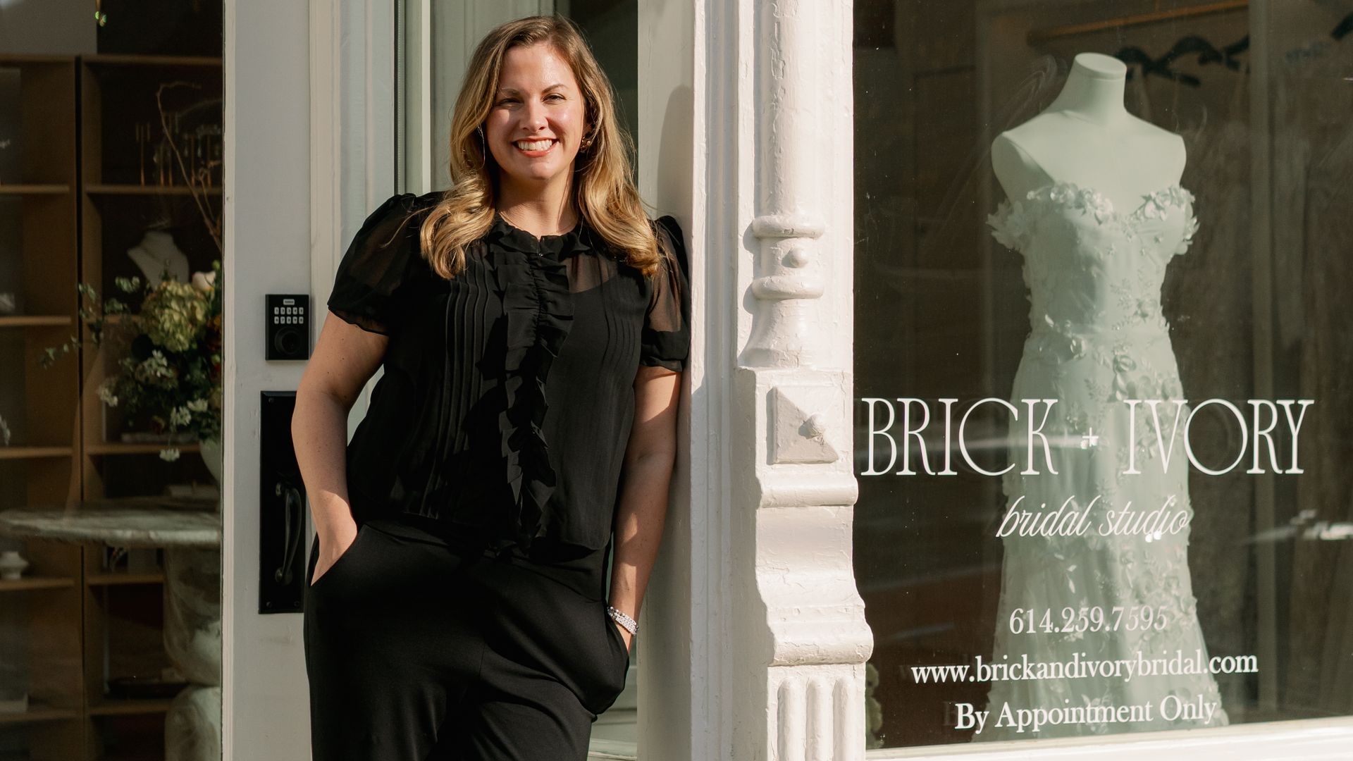 Woman in black outfit smiling and leaning against white storefront corner of Brick + Ivory bridal studio with a white wedding dress mannequin visible through the window.