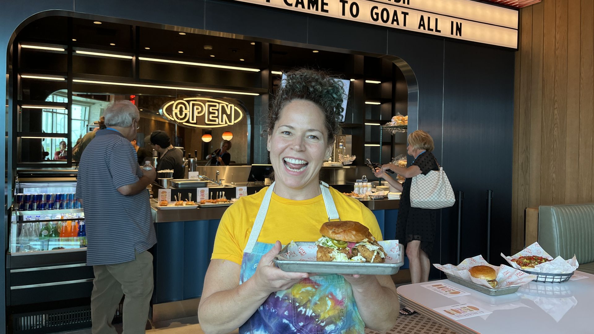 A smiling woman in a yellow shirt and colorful apron holds a sandwich on a tray inside a food shop with an "OPEN" neon sign and a marquee reading "I DIDN'T COME TO PLAY SHEEPISH, I CAME TO GOAT ALL IN".