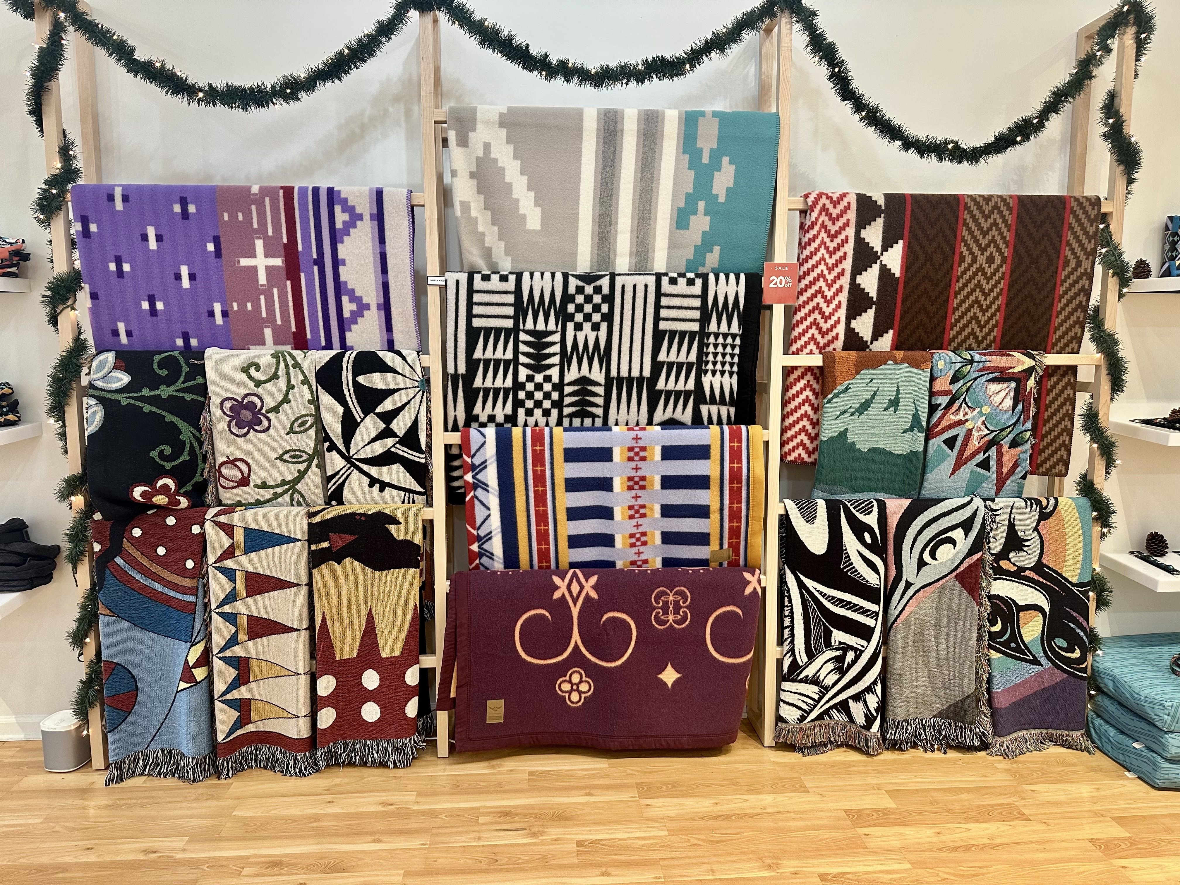 Display of colorful woven blankets hung on wooden racks, featuring geometric and floral patterns in purple, gray, teal, black, red, and brown. Garlands drape overhead.