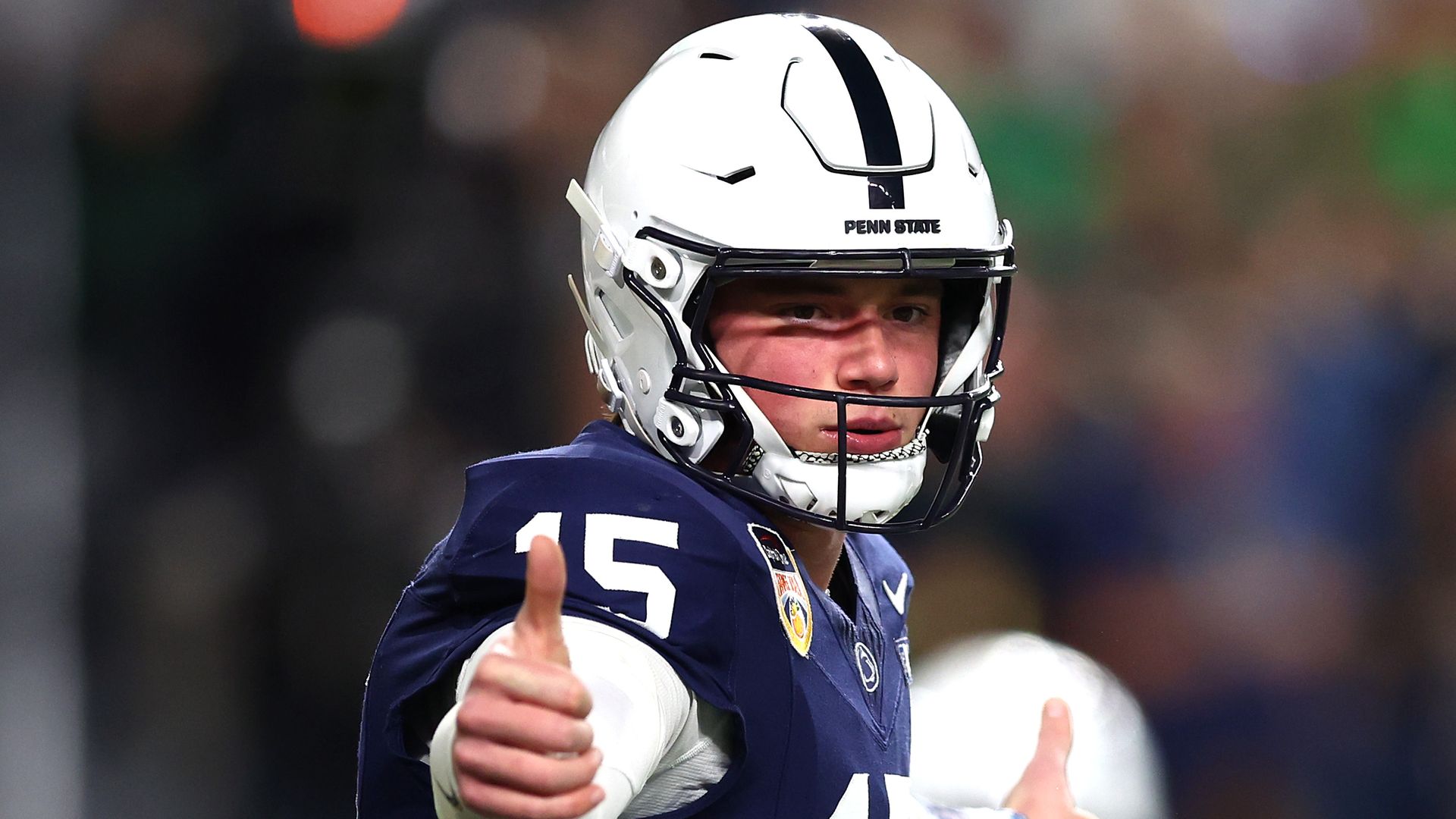 Penn State football player in navy blue jersey number 15 and white helmet giving thumbs up during a game, blurred crowd in background.