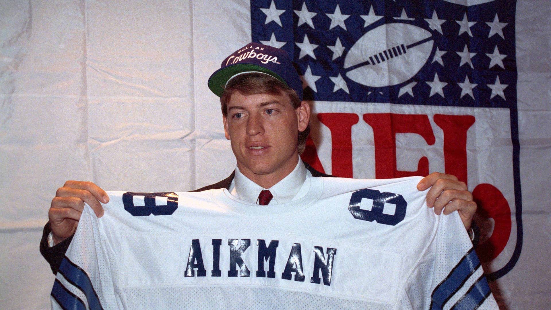 A man wearing a Dallas Cowboys baseball cap holds up a jersey saying "AIKMAN"