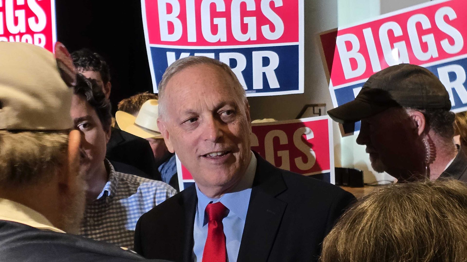 Andy Biggs in a dark suit and red tie speaks with supporters at a rally, surrounded by bright red, white, and blue campaign signs reading "BIGGS/KERR."