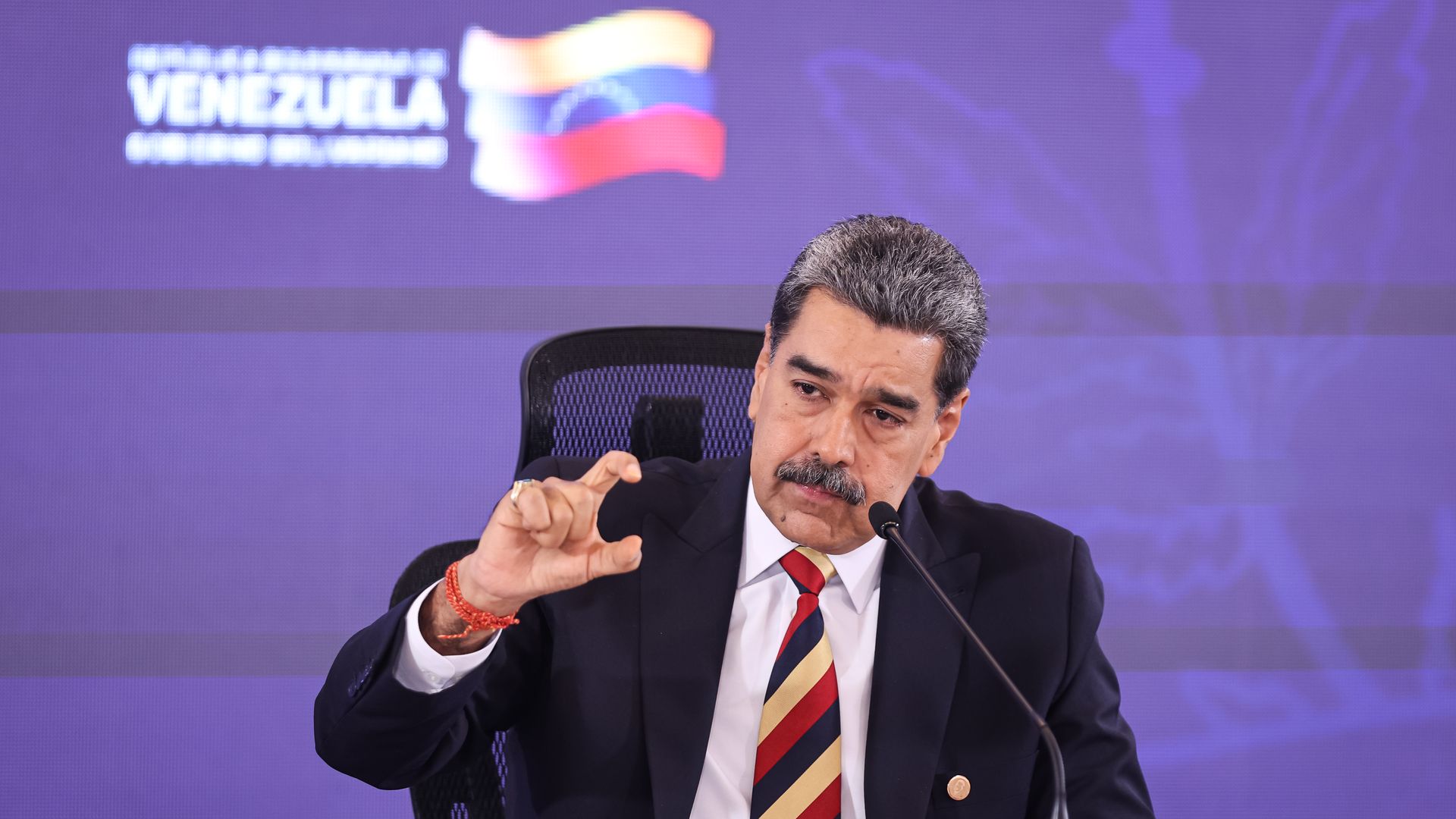 Venezuelan leader Nicolás Maduro, with graying black hair and wearing a red, blue and gold striped tie, a white shirt and navy jacket, makes his thumb and index finger into a "c" shape in front of a purple background featuring the Venezuelan flag and the word "VENEZUELA" in white.