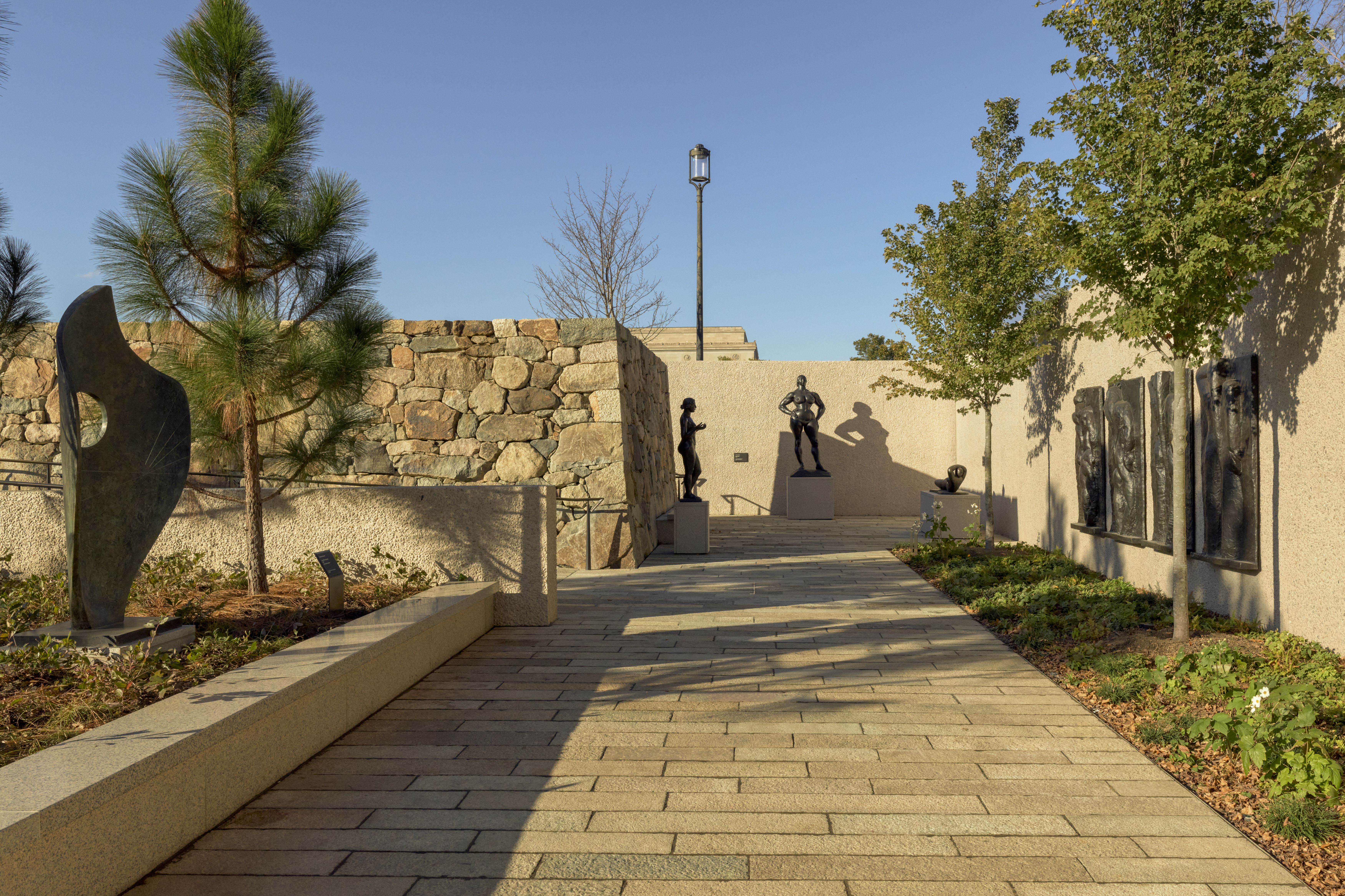 Sunny outdoor sculpture garden with a beige stone wall, paved path, and a tall lamp. A central pedestal statue of a muscular nude man is flanked by smaller figures; trees frame the scene.