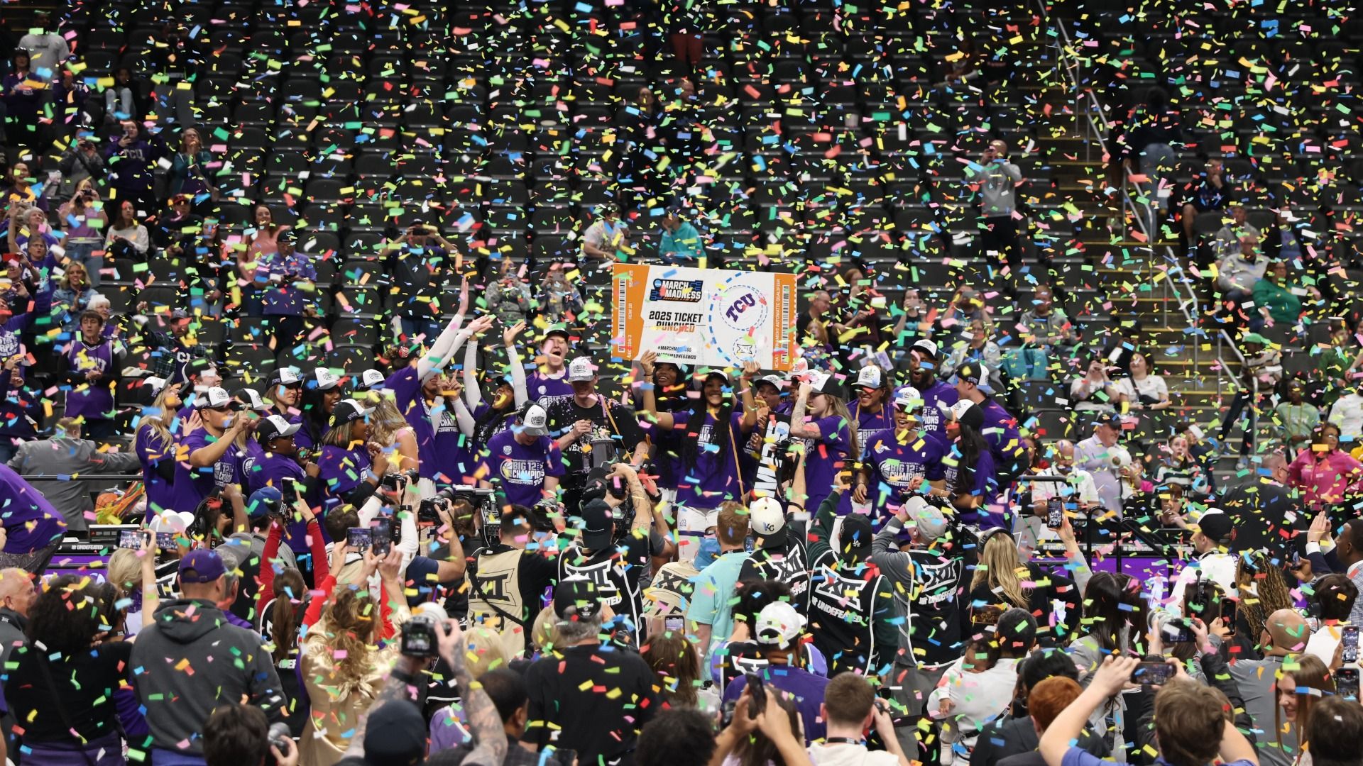 Celebrating sports team in purple uniforms holding a large March Madness 2025 ticket sign amid falling multicolored confetti with crowd and photographers capturing the moment.