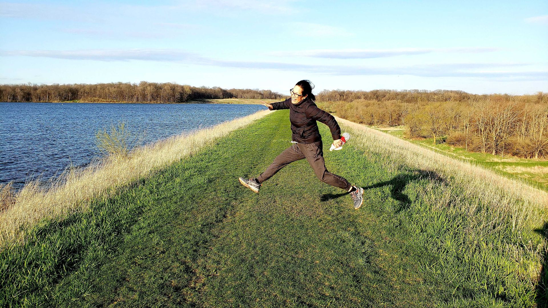 Photo of a woman jumping up and down at a park next to a pond. 