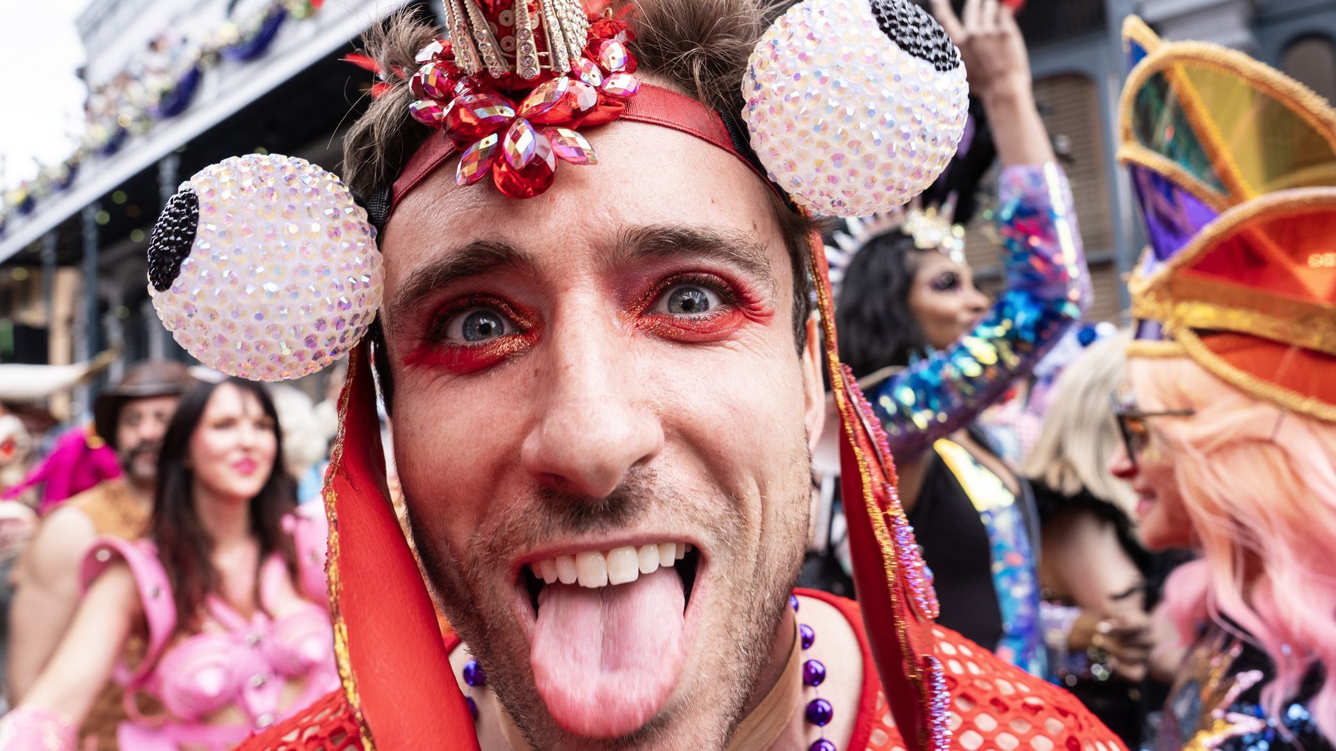 Man wearing a red feathered headdress with large glittery eyeball decorations, red eye makeup, sticking his tongue out at a colorful costume party or parade, with other costumed people in the background.