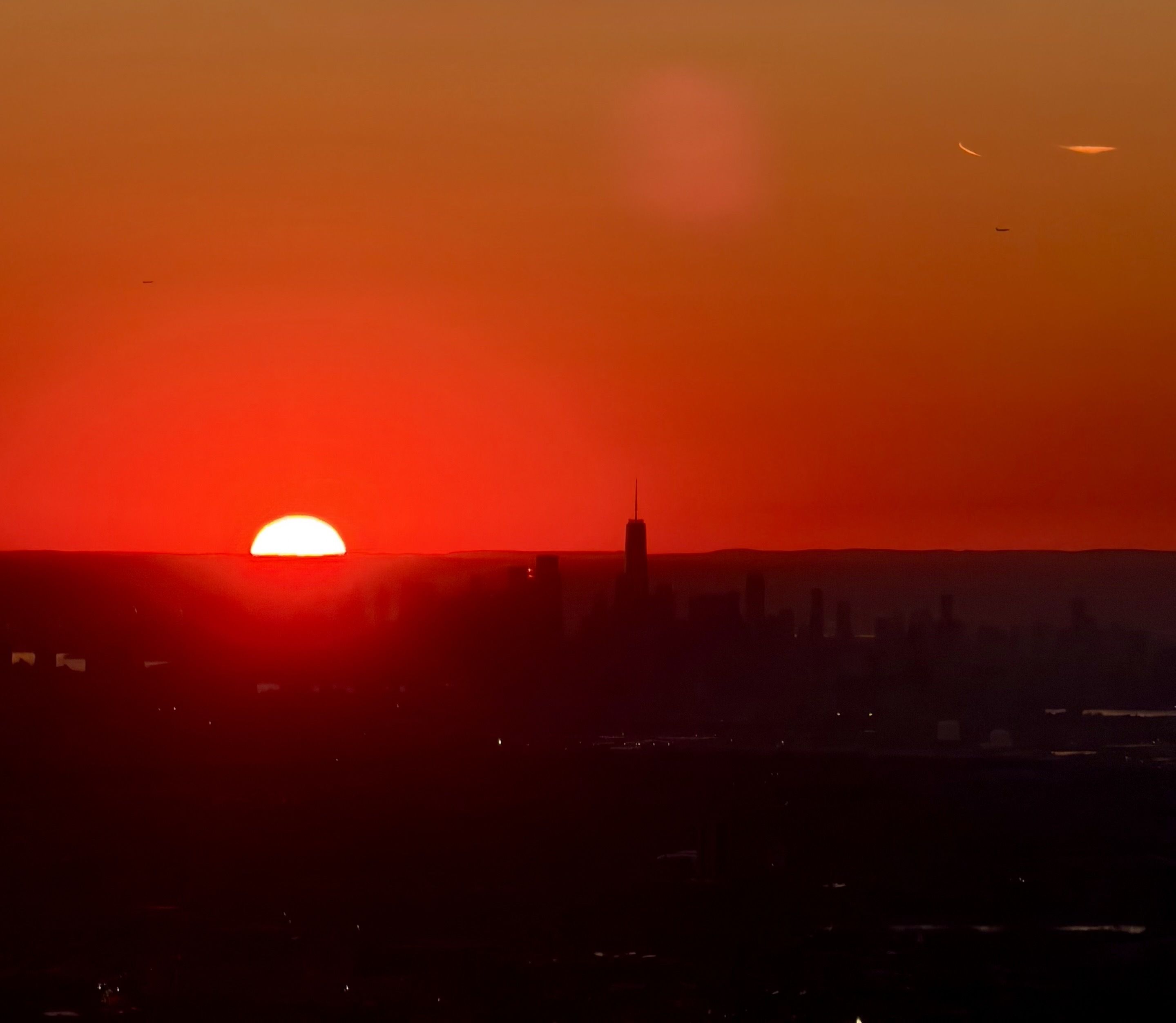Silhouetted city skyline at sunset with a bright red sun half-visible on the horizon; a tall central tower rises above shorter buildings, against an orange and red sky.