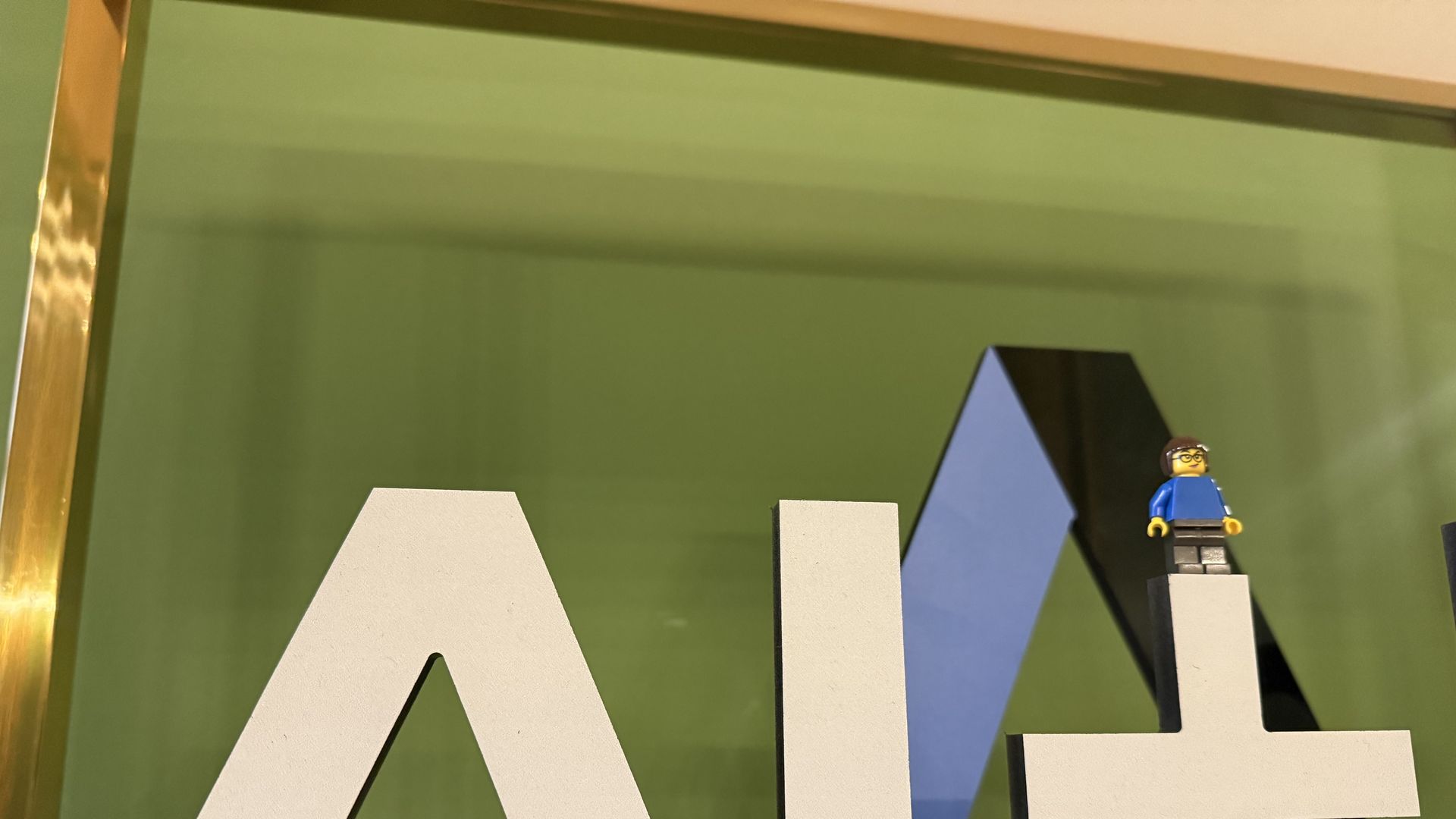 White block letters "AI+" on a black base, with a small Lego figure wearing a blue shirt and glasses standing on the plus sign, against a green background with gold trim.