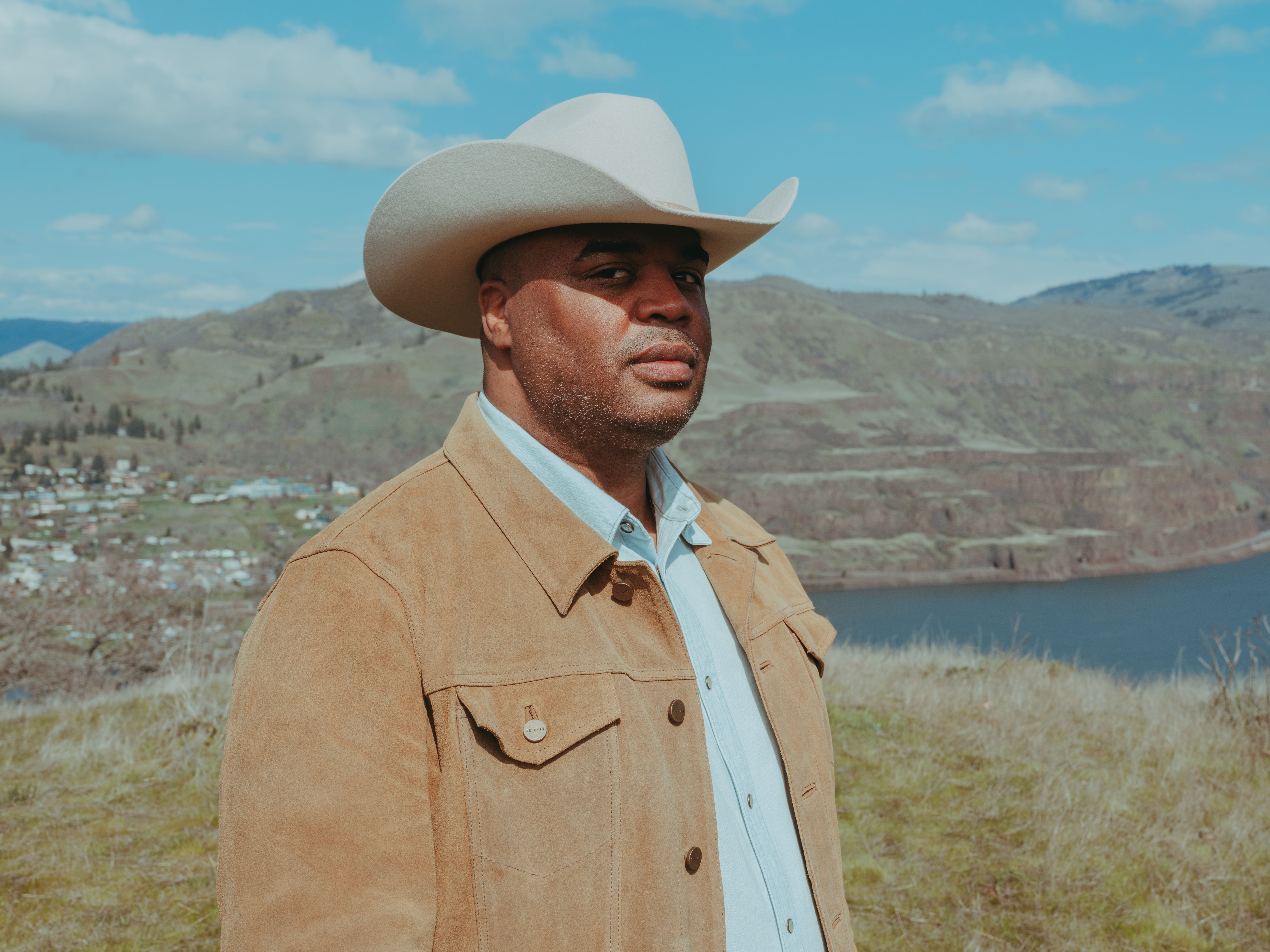 A Black man wearing a white cowboy hat, tan jacket and light blue shirt stands above a deep river gorge on a sunny day.
