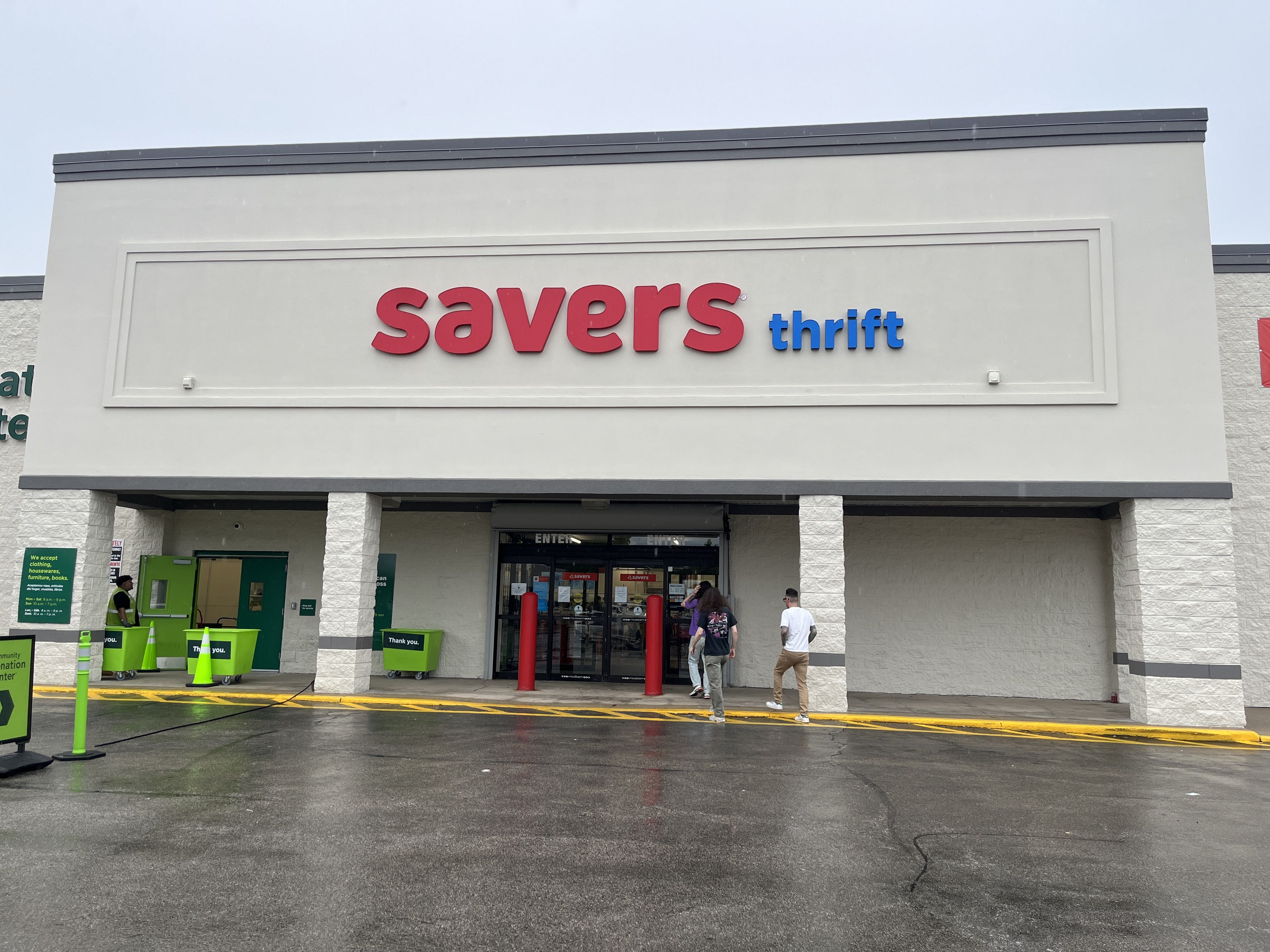 Exterior of a thrift store with a large red savers and blue thrift sign on a beige wall; glass entrance doors, green shopping carts outside, and wet pavement.