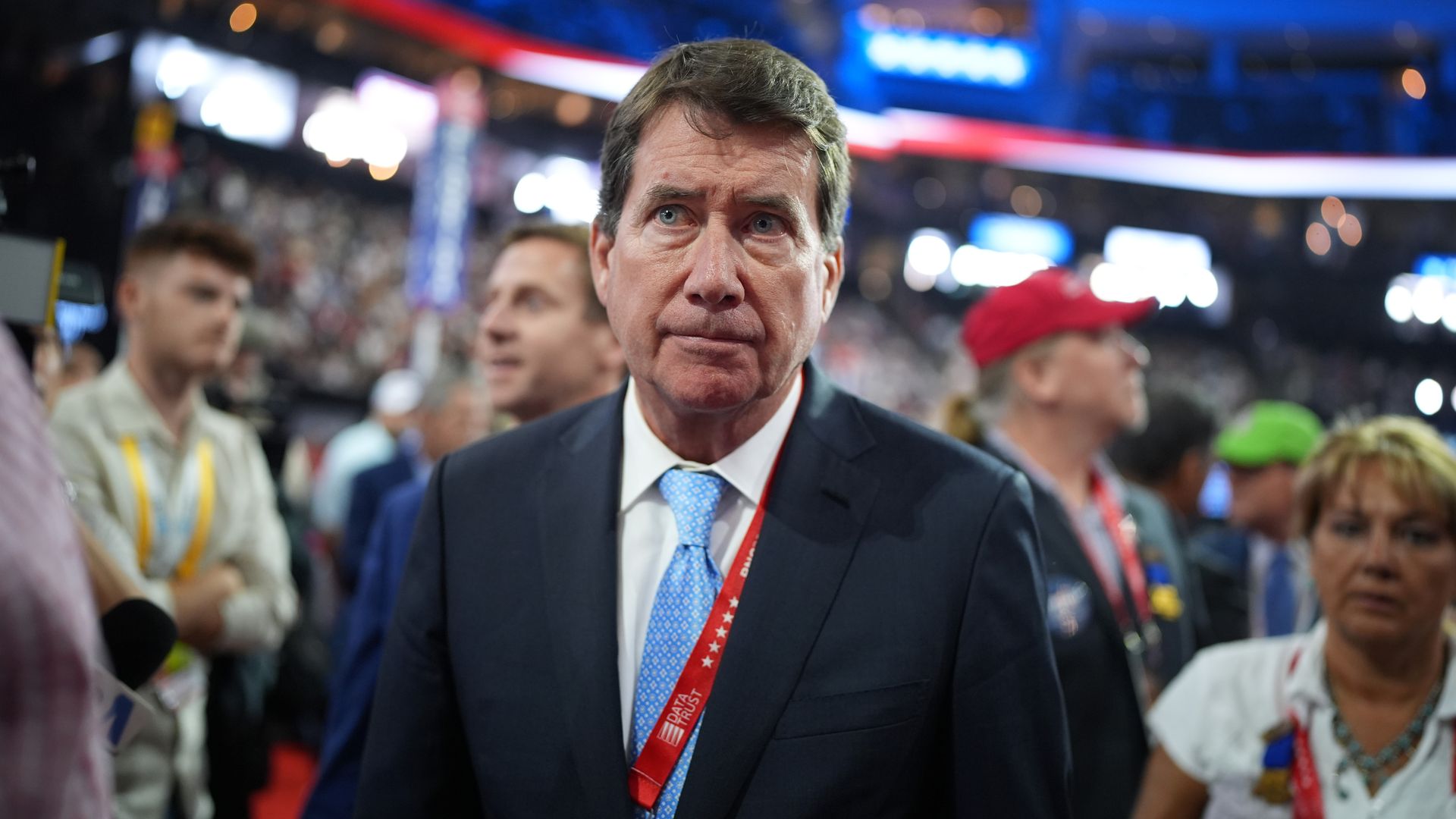 Portrait of a distinguished man in a blue business suit with a light blue tie in a busy room, with many people behind him. Lots of red, white and blue in the background.