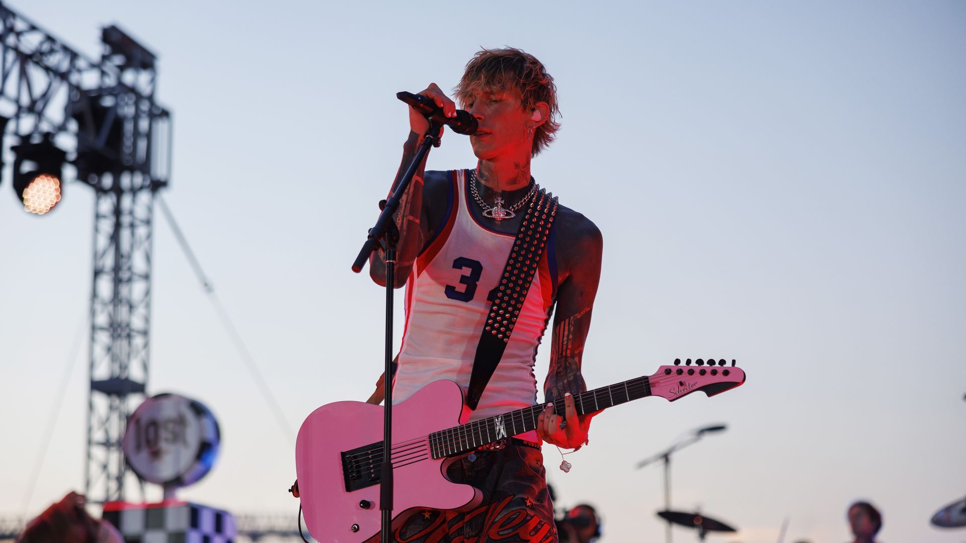 Outdoor concert: tattooed blond guitarist sings into a mic while playing a pink electric guitar. He wears a white tank top with the number 3 and a studded strap, red lights on his face.