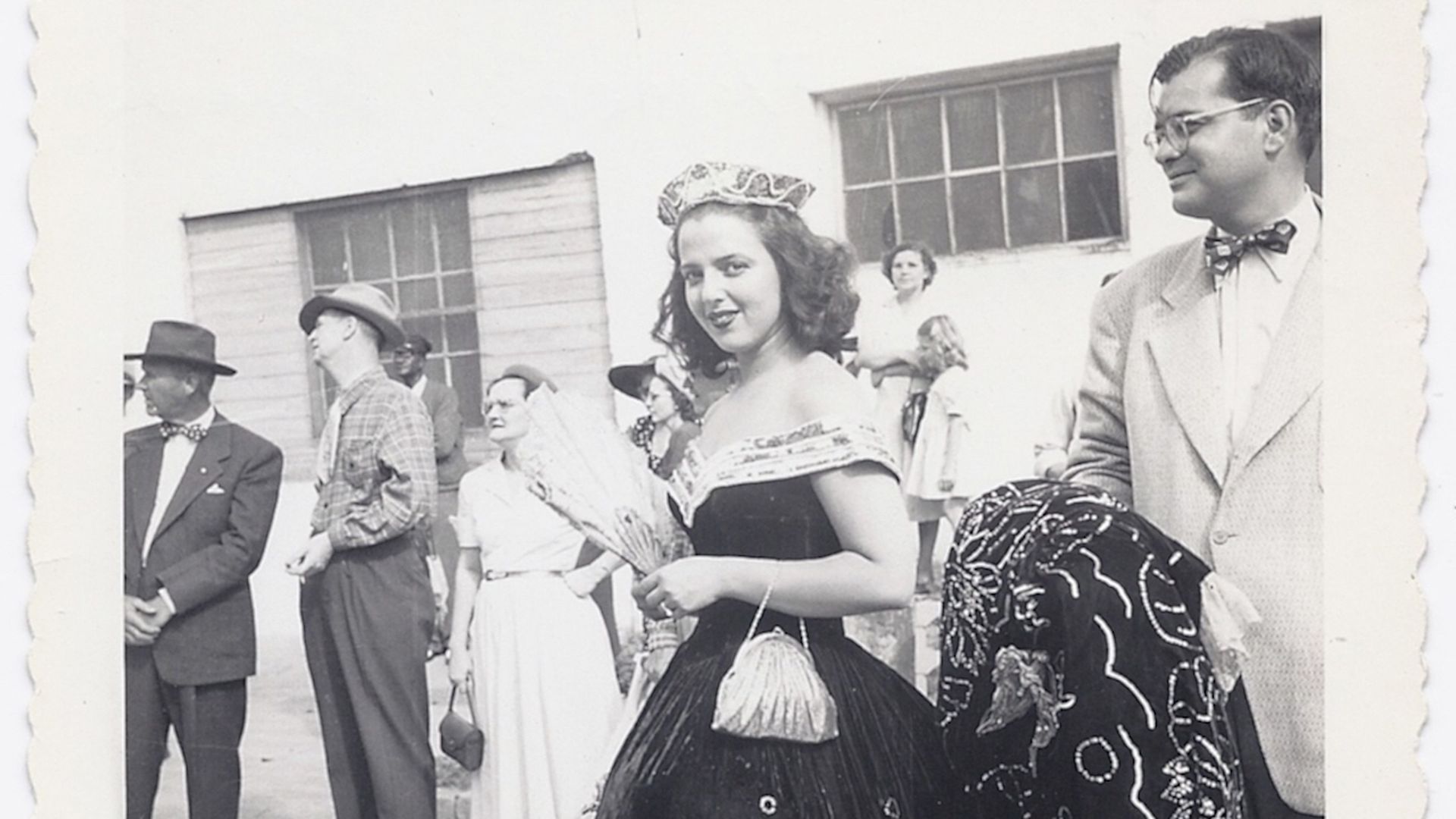 A woman in a regal dress with a fan and a crown in a black and white photo.