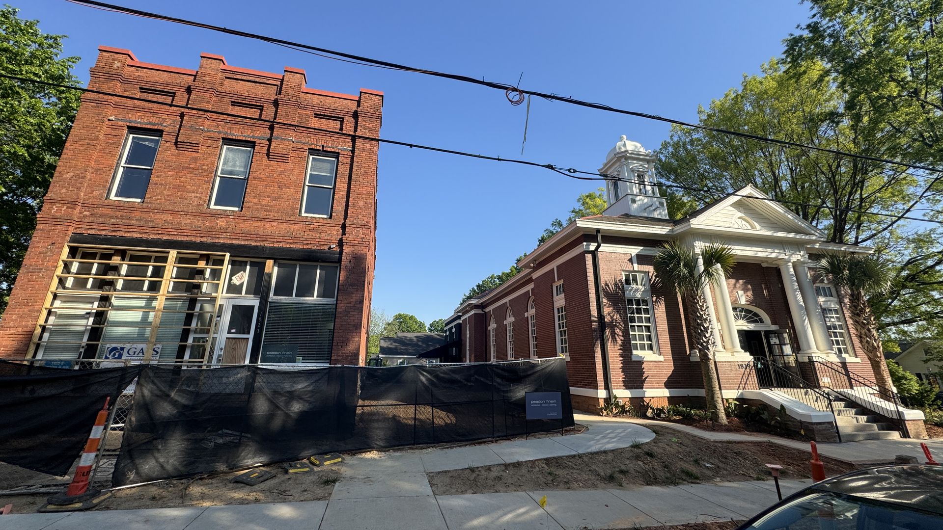 Two adjacent brick buildings under clear blue sky; left building is older with boarded windows and construction fencing, right building has white columns and a small tower, surrounded by trees.