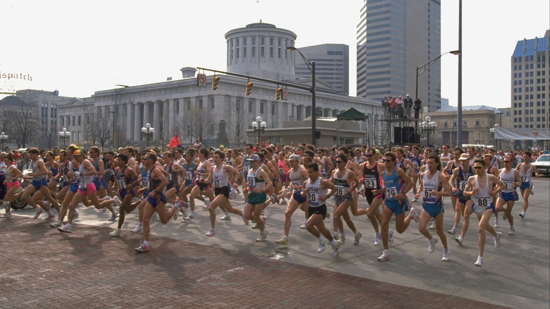 Dozens of marathon runners run through an intersection in downtown Columbus with the Ohio Statehouse in the background. 