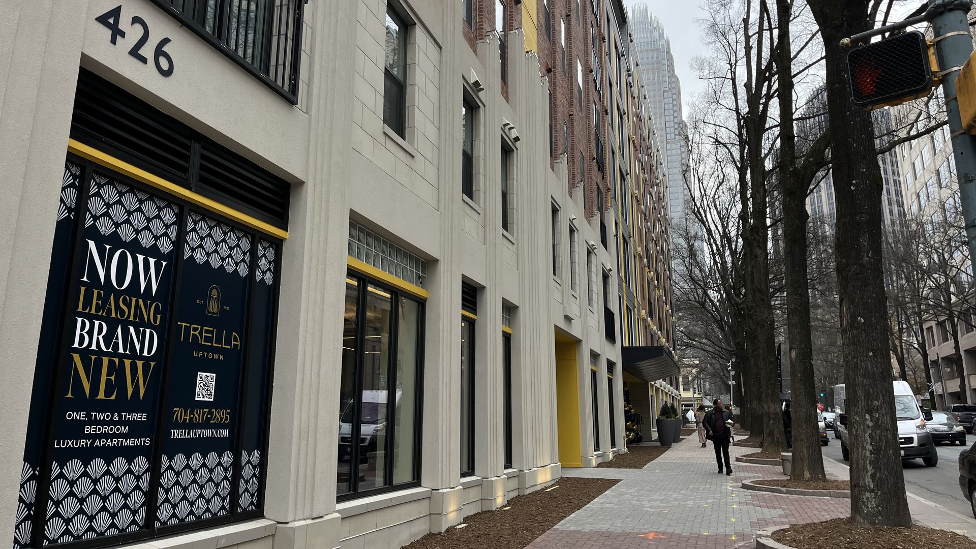 Street view of a modern apartment building at 426 with a "Now Leasing Brand New" sign in black, gold, and white. Sidewalk lined with trees and a few pedestrians under a gray sky.