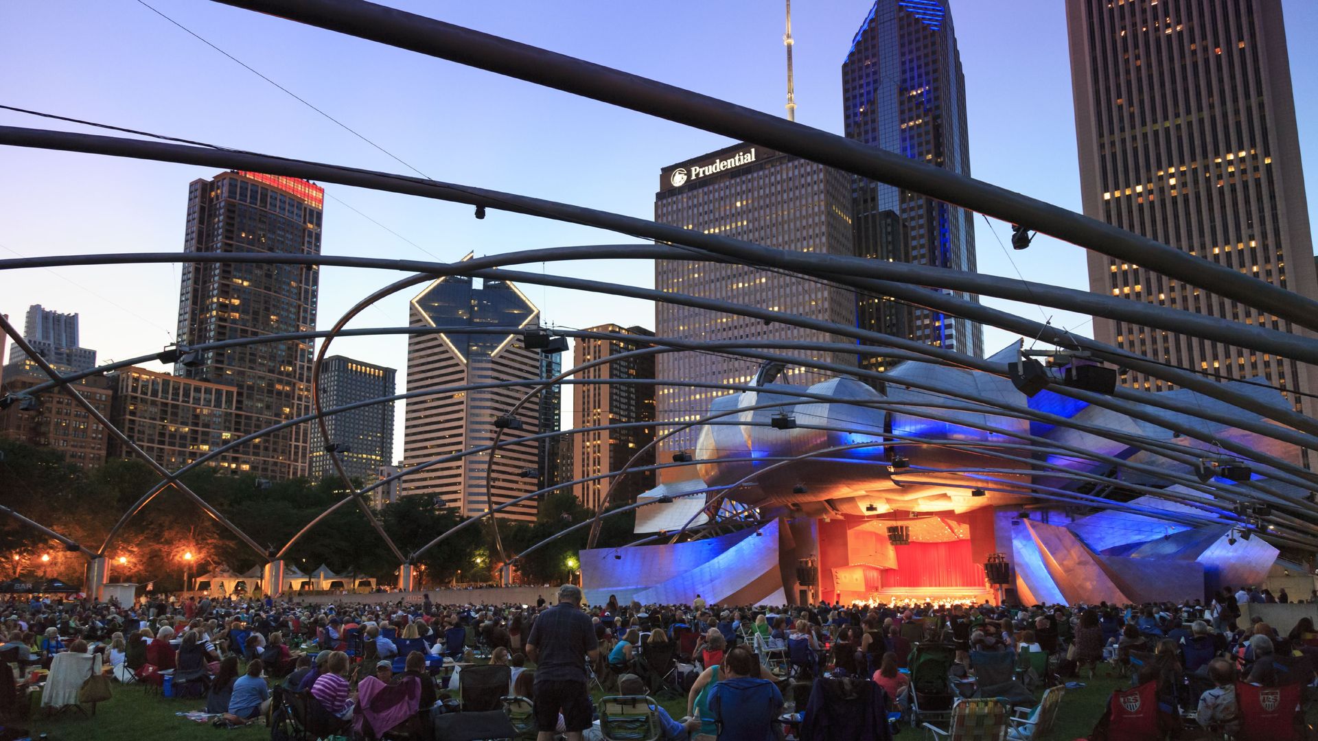 Crowd gathered at Jay Pritzker Pavilion in Chicago's Millennium Park during evening event, with illuminated silver stage and city skyscrapers, including Prudential building, in the background.