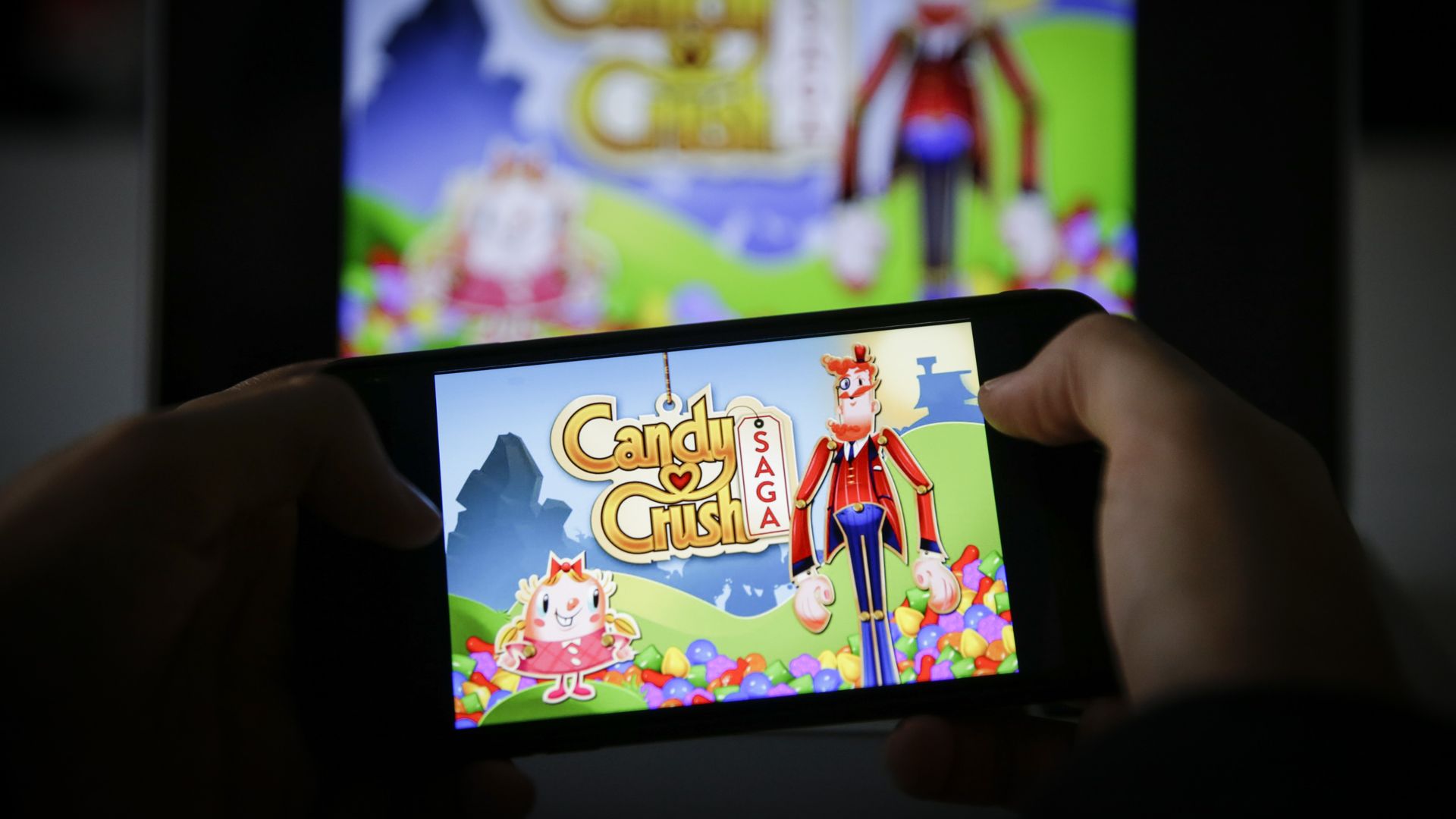 In this image, someone holds a phone that displays the Candy Crush home page in front of a TV screen that also displays Candy Crush.