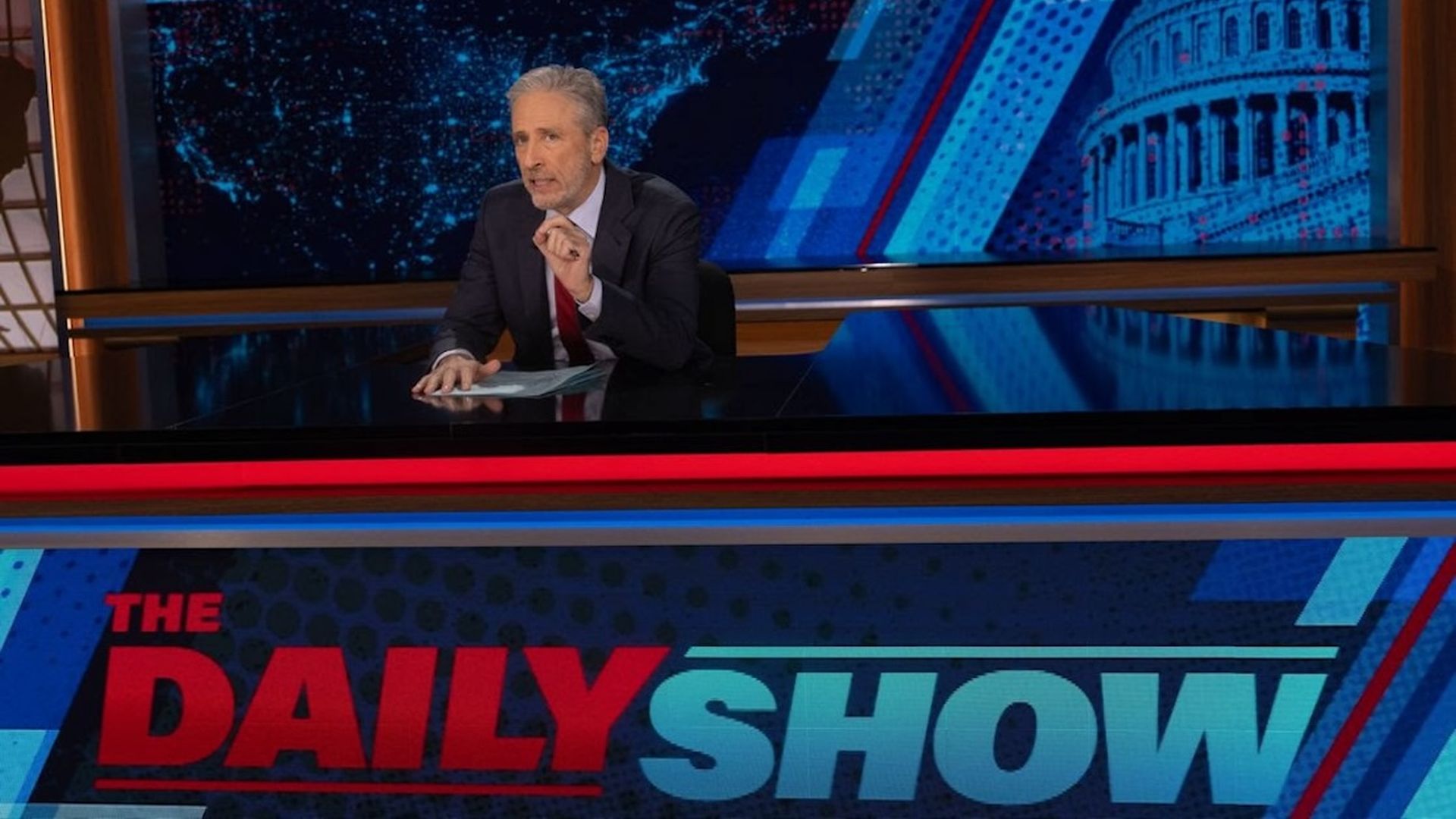 Jon Stewart sits behind the Daily Show desk.