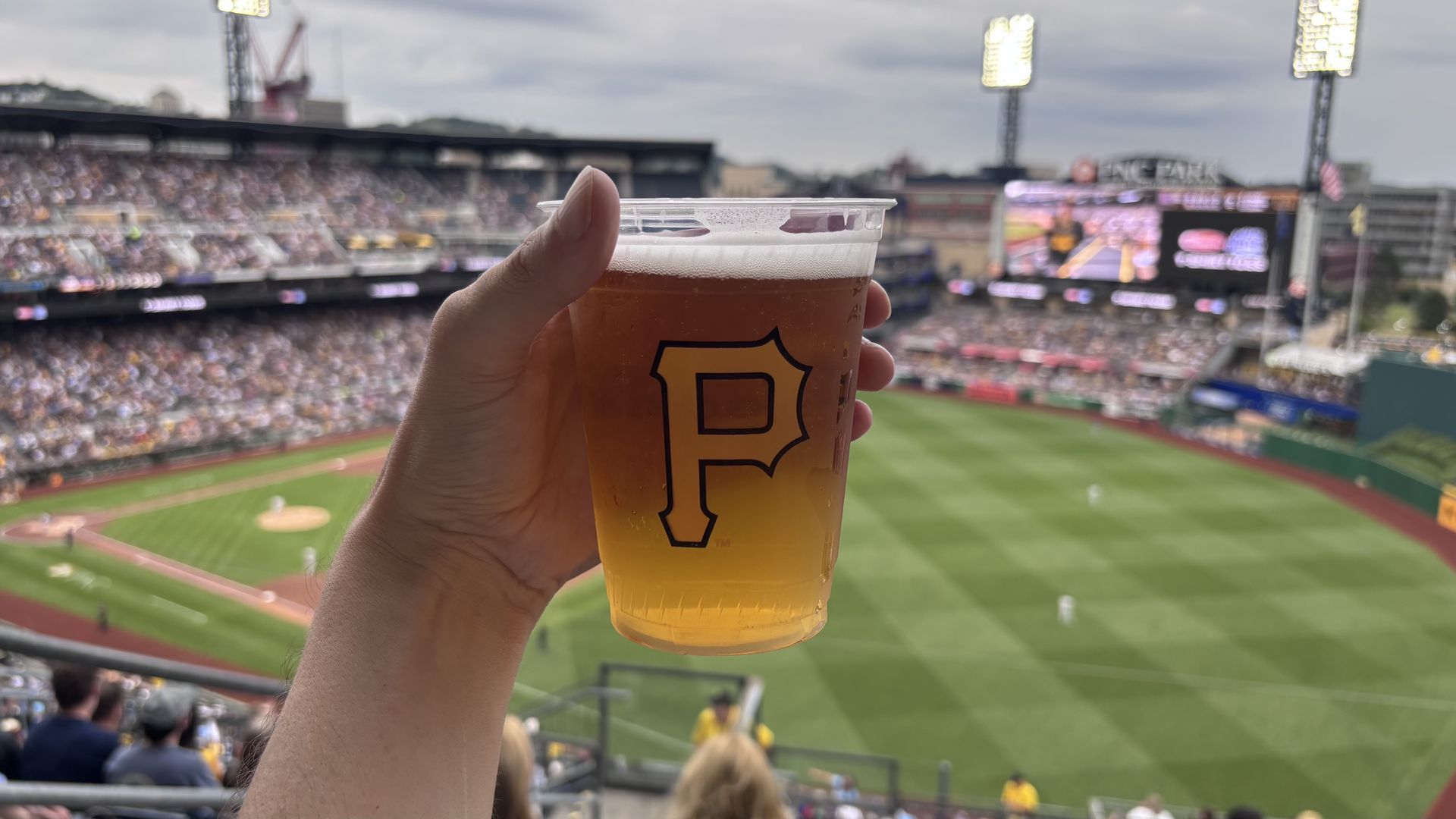 A hand holds a clear plastic beer cup with a large black and yellow logo, set against a crowded baseball stadium with a green field and a large scoreboard in the background.