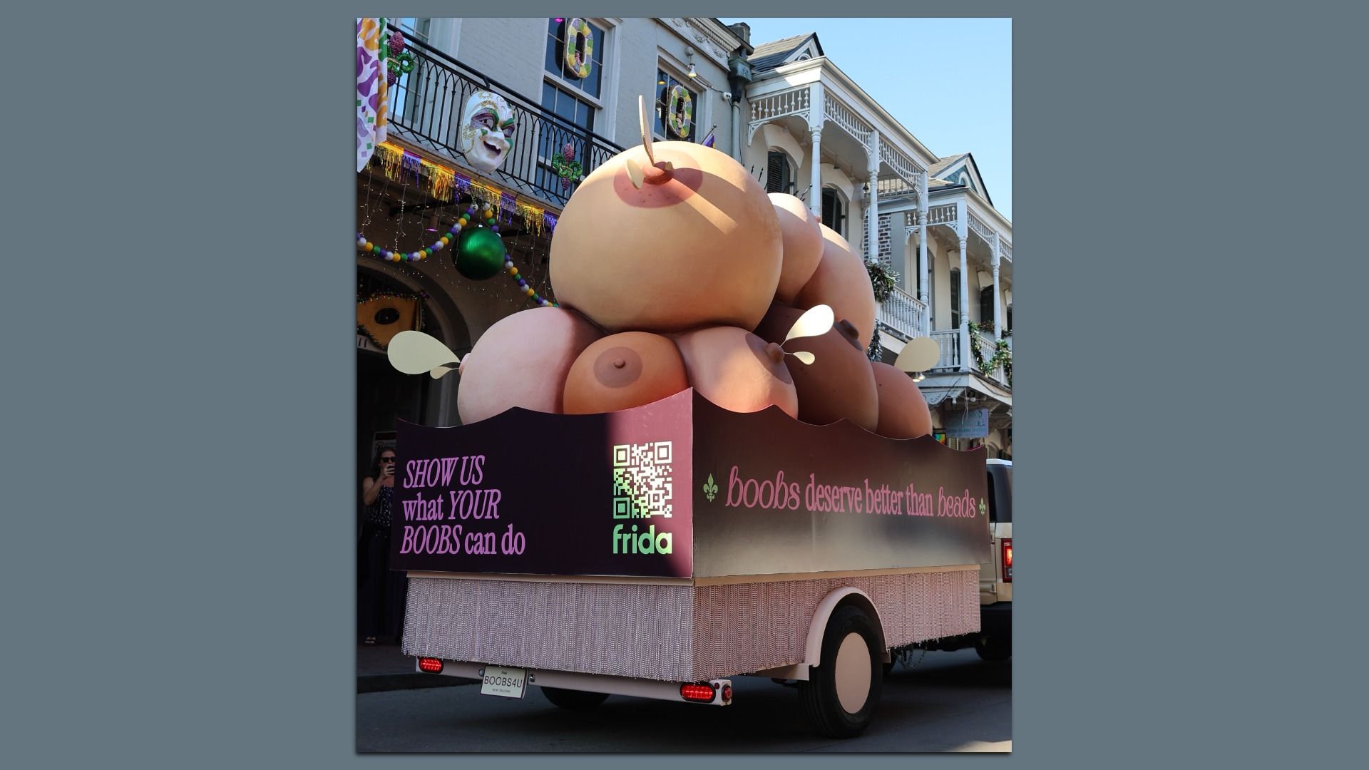 Float with large, varied breast sculptures on a vehicle, displaying messages "SHOW US what YOUR BOOBS can do" and "boobs deserve better than beads" in pink text, festive street background.