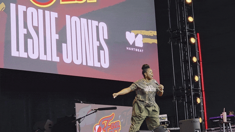 Leslie Jones performing on stage in green pants and a black "God's Favorite" t-shirt, with her name in large white letters on a screen behind her at the Hartbeat event.