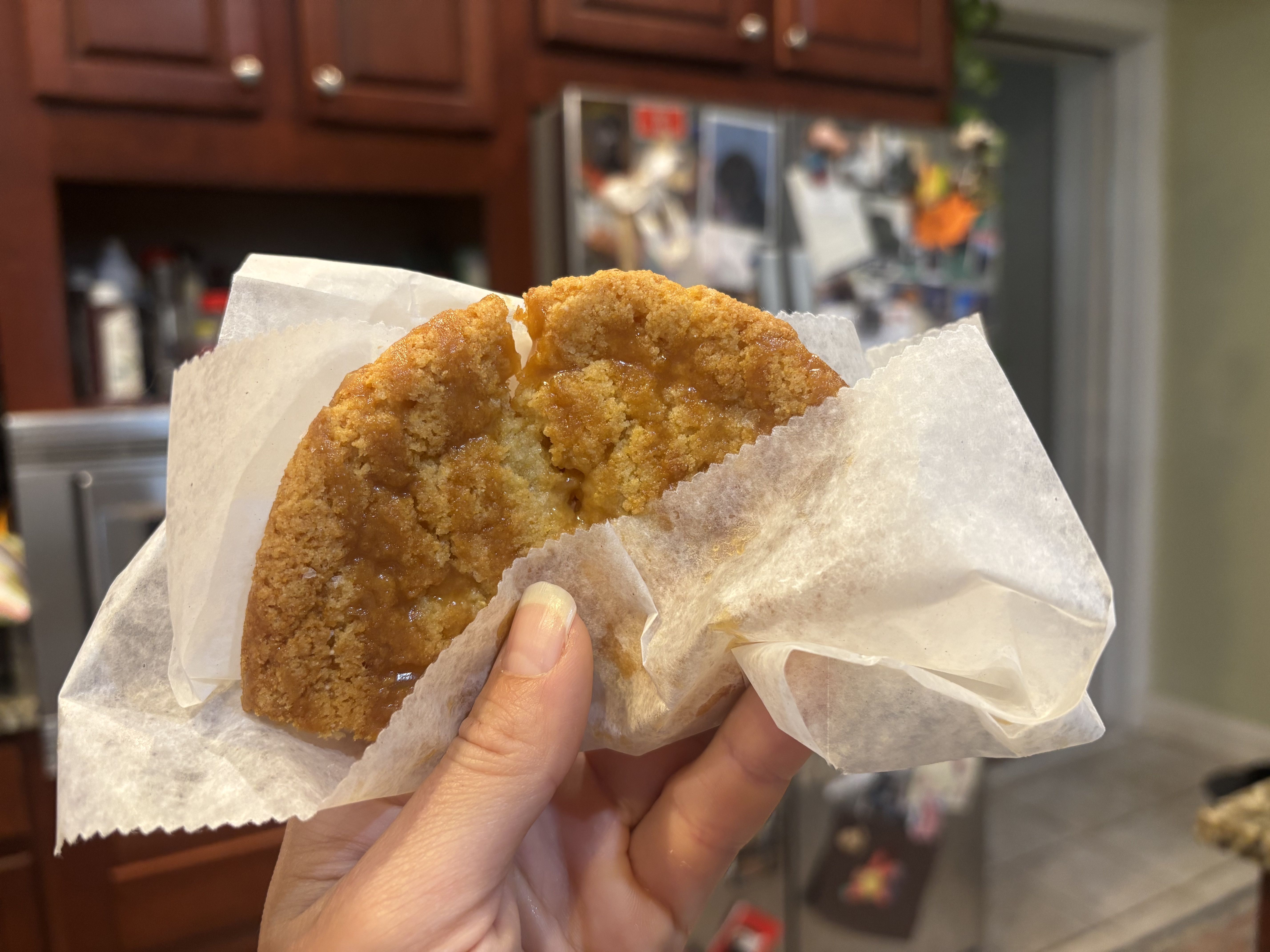A hand holding a crumbly, caramel-glazed fried pastry in a white paper bag, with a blurred kitchen and refrigerator in the background.
