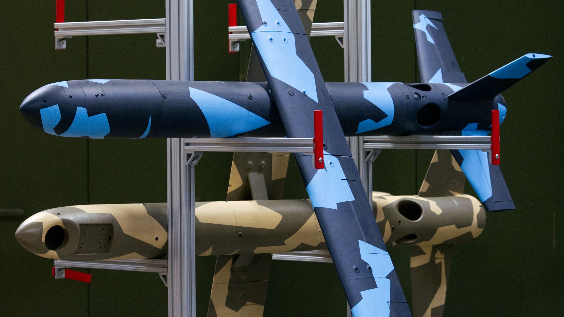 Two drones, one in desert camo and one in arctic camo, sit on a rack inside a factory.