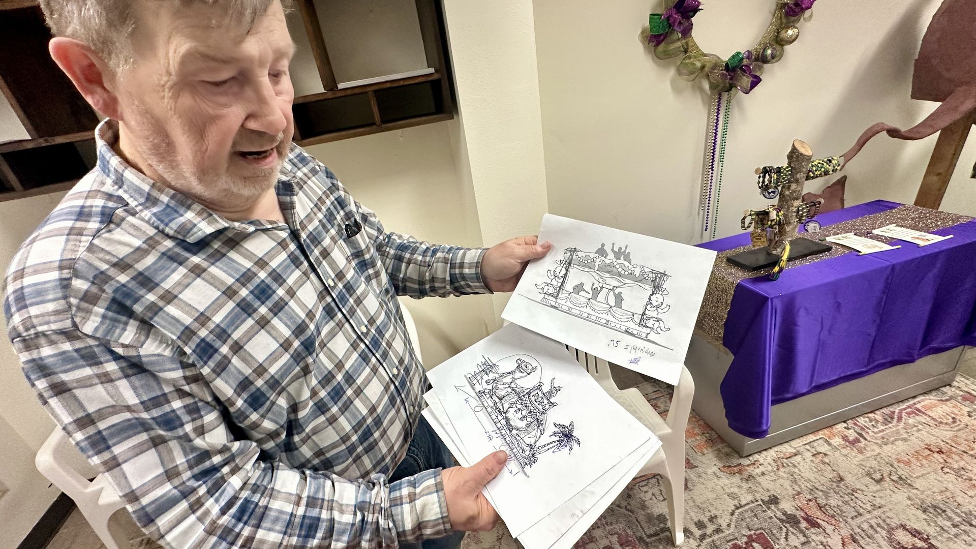A man in a blue and white plaid shirt holding black-and-white sketches, standing next to a table with a purple cloth and a Mardi Gras-themed wreath and jewelry display.