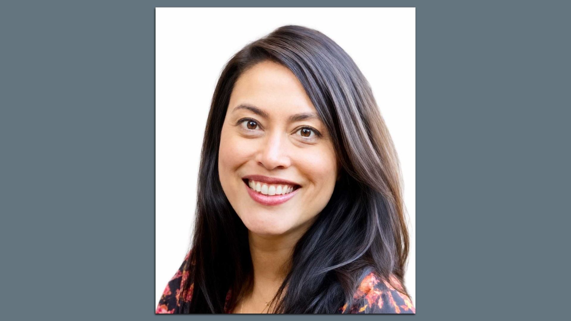Close-up portrait of a smiling woman with long dark hair, light makeup, and brown eyes, wearing a floral top, against a white background.
