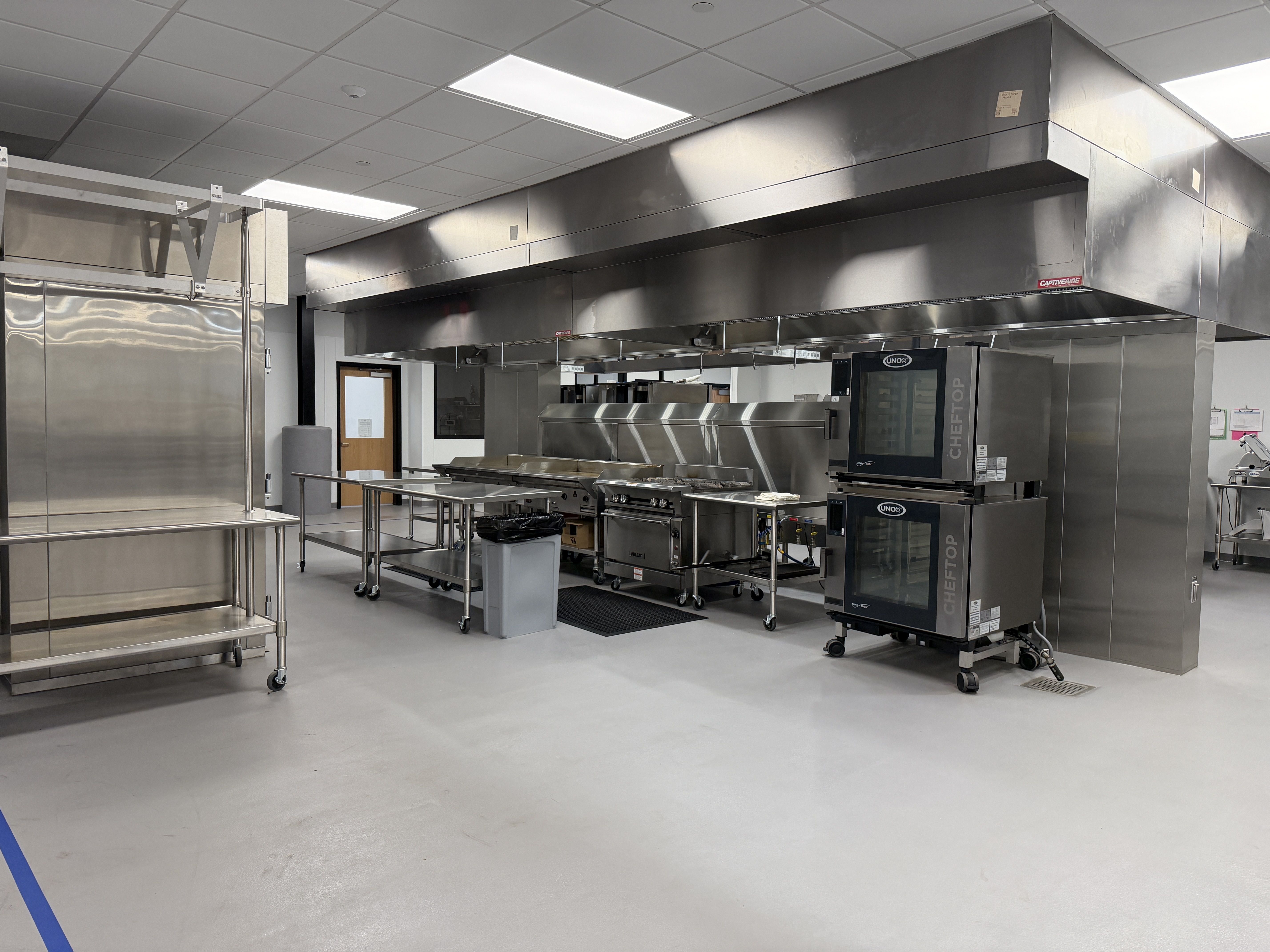 Industrial kitchen with stainless steel appliances and large overhead exhaust hoods. On wheels prep tables, bright white lighting, and a blue floor line marking the area.